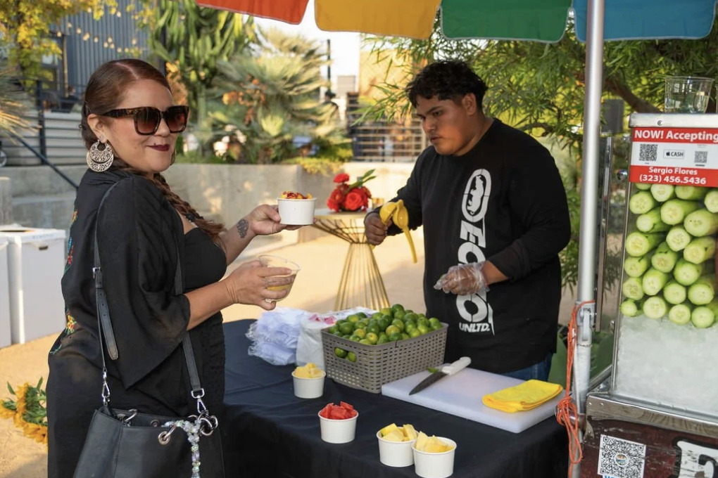 A woman with sunglasses and earrings is holding a cup of drink and a bowl of food while standing at a fruit stand with a young man preparing lime and fruit drinks at an outdoor market.
