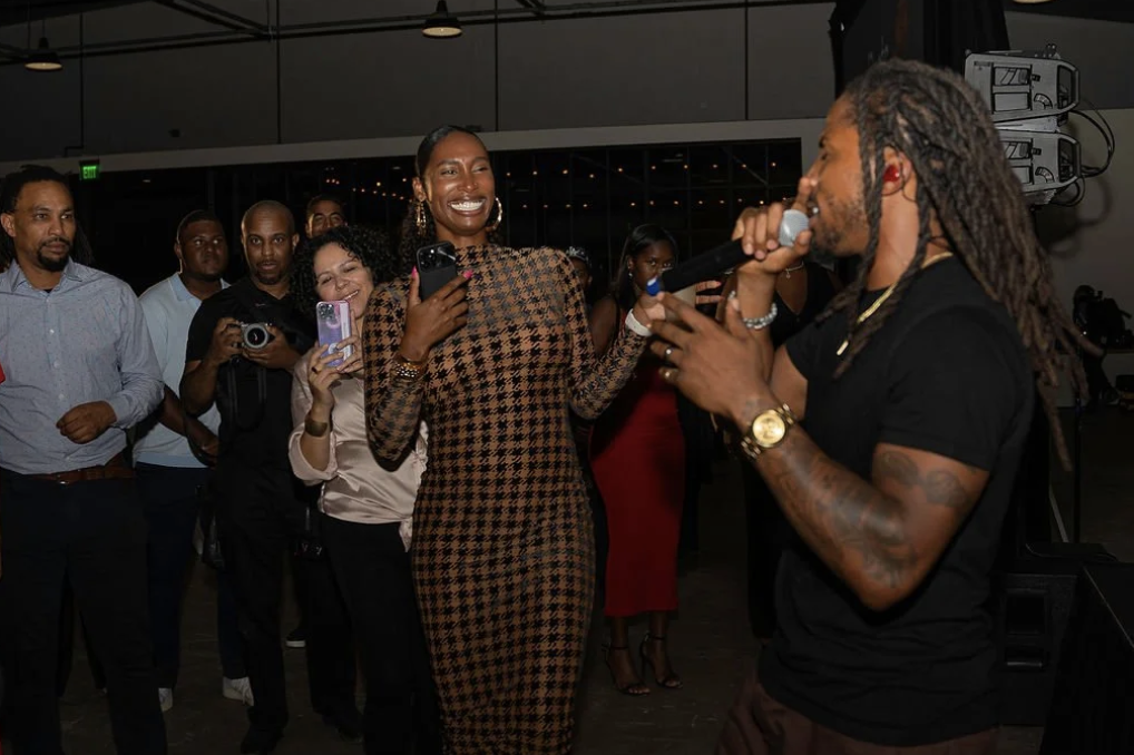 A group of people at an indoor event, with one woman in a houndstooth dress smiling while engaging with a man holding a microphone, who has long dreadlocks and tattoos.