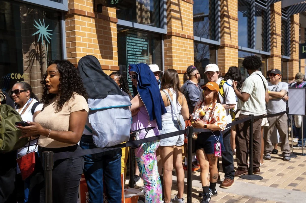 Line of diverse people waiting outside a building with large windows and brick facade on a sunny day.