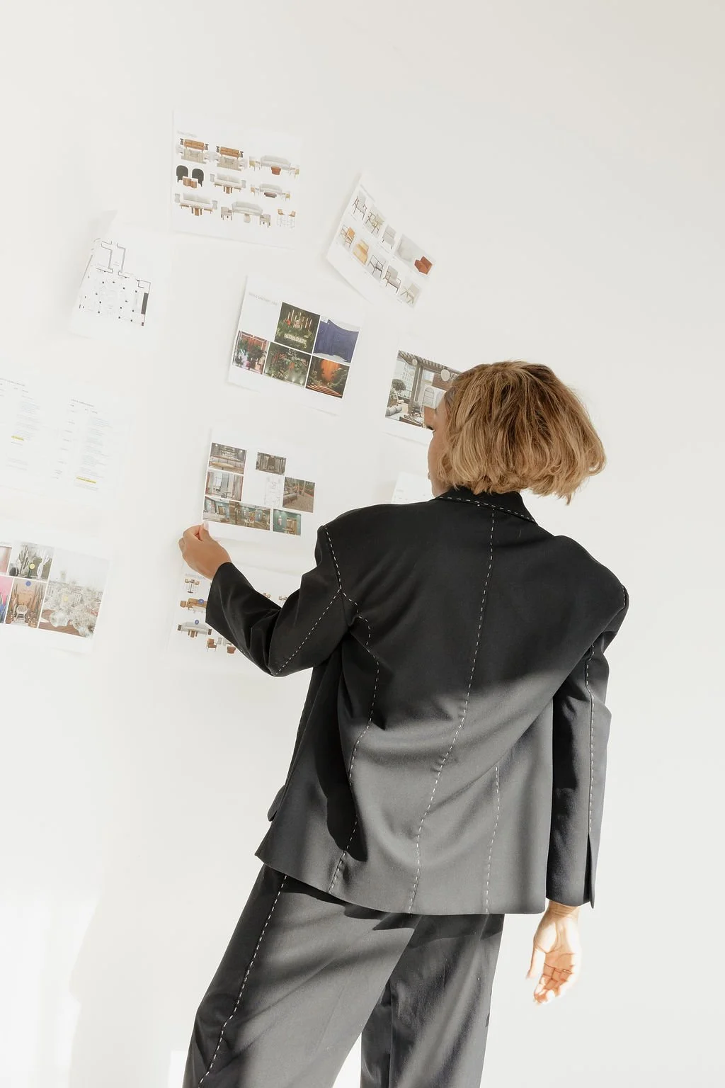 Person with short blonde hair wearing a black suit looking at images and plans on a white wall.