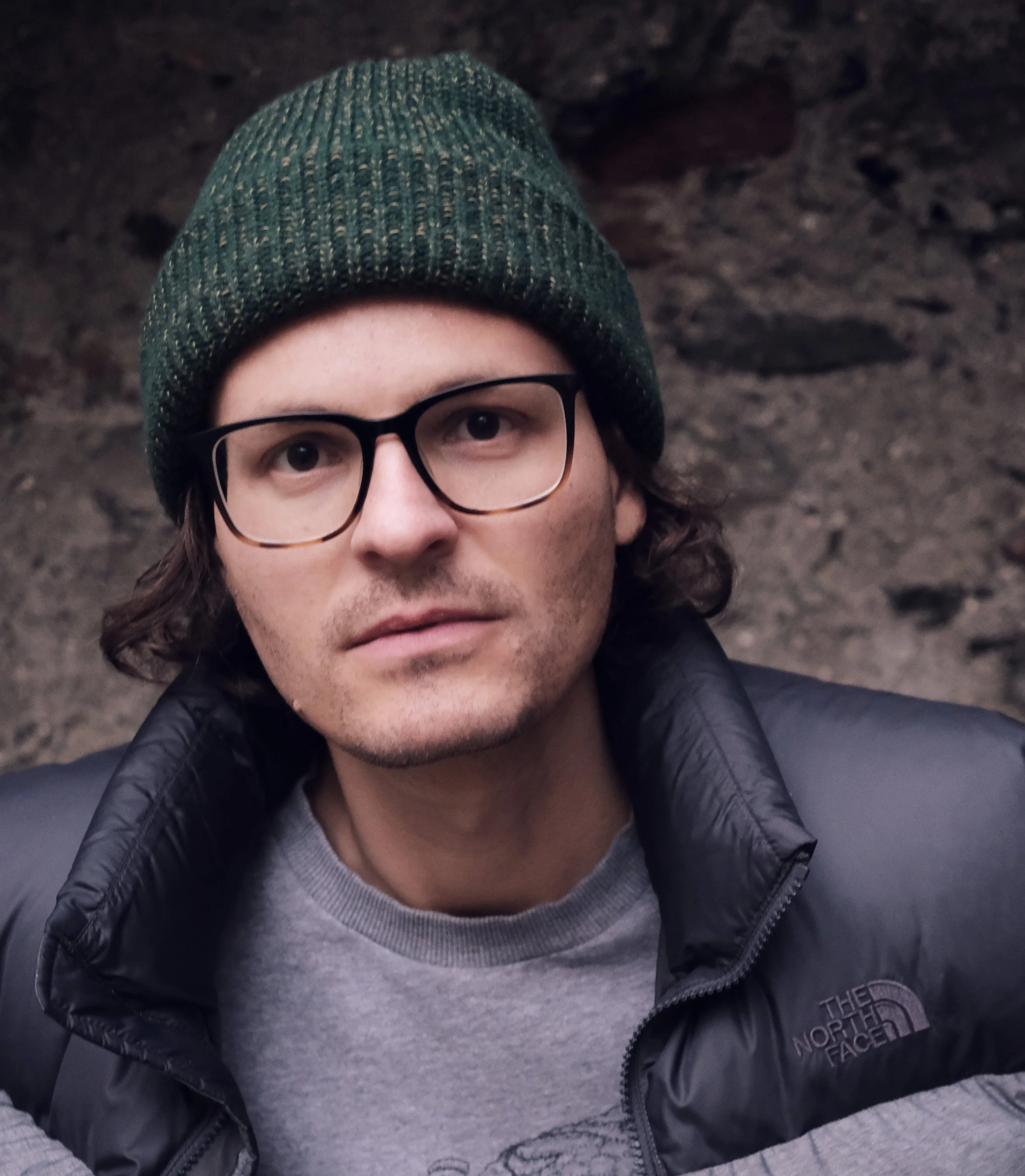 Close-up photo of a young man with glasses, wearing a green knit beanie and a black North Face jacket, against a background of dark rocky terrain.
