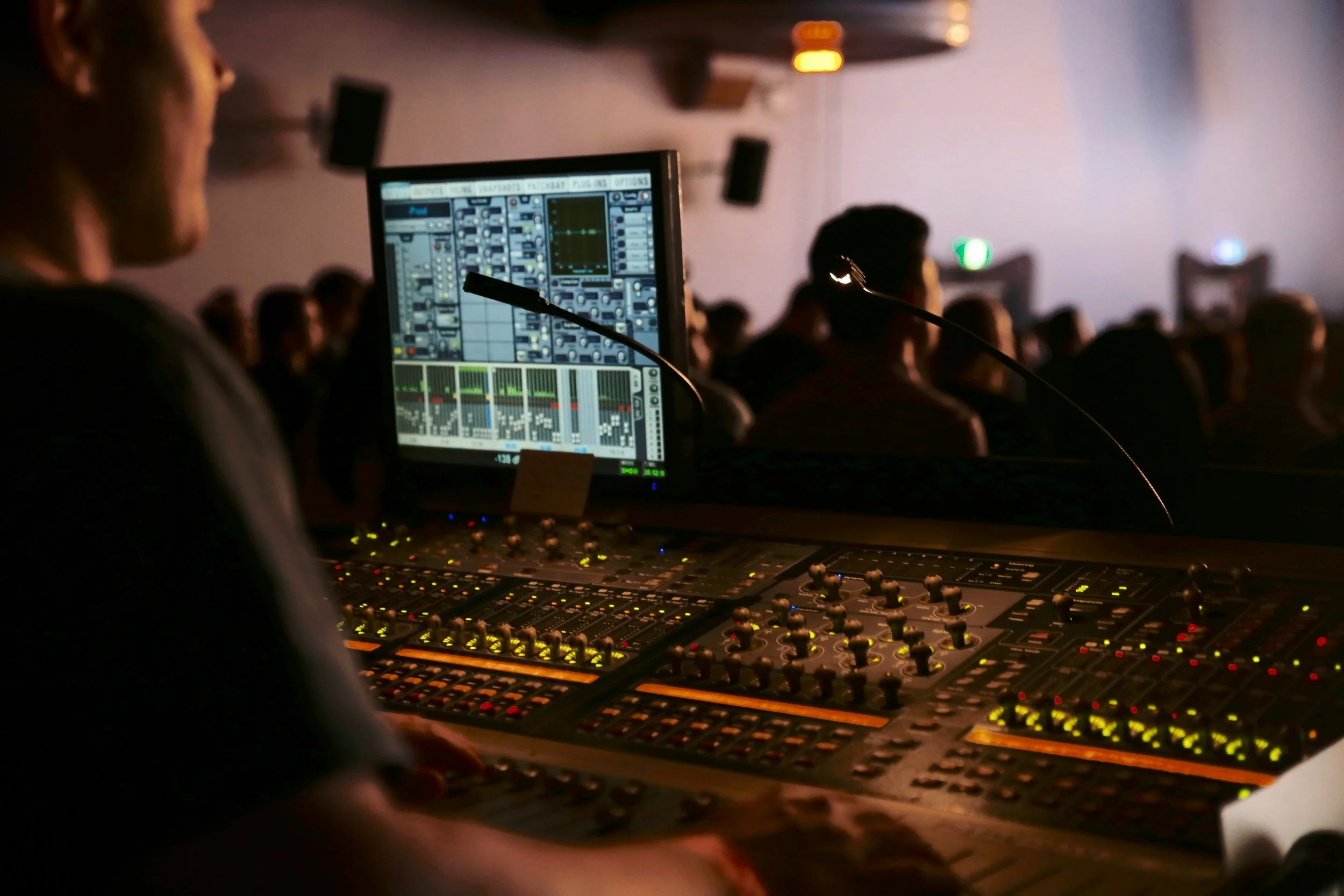 A person operating an audio mixing console with a computer screen displaying sound controls in a dimly lit auditorium filled with an audience.