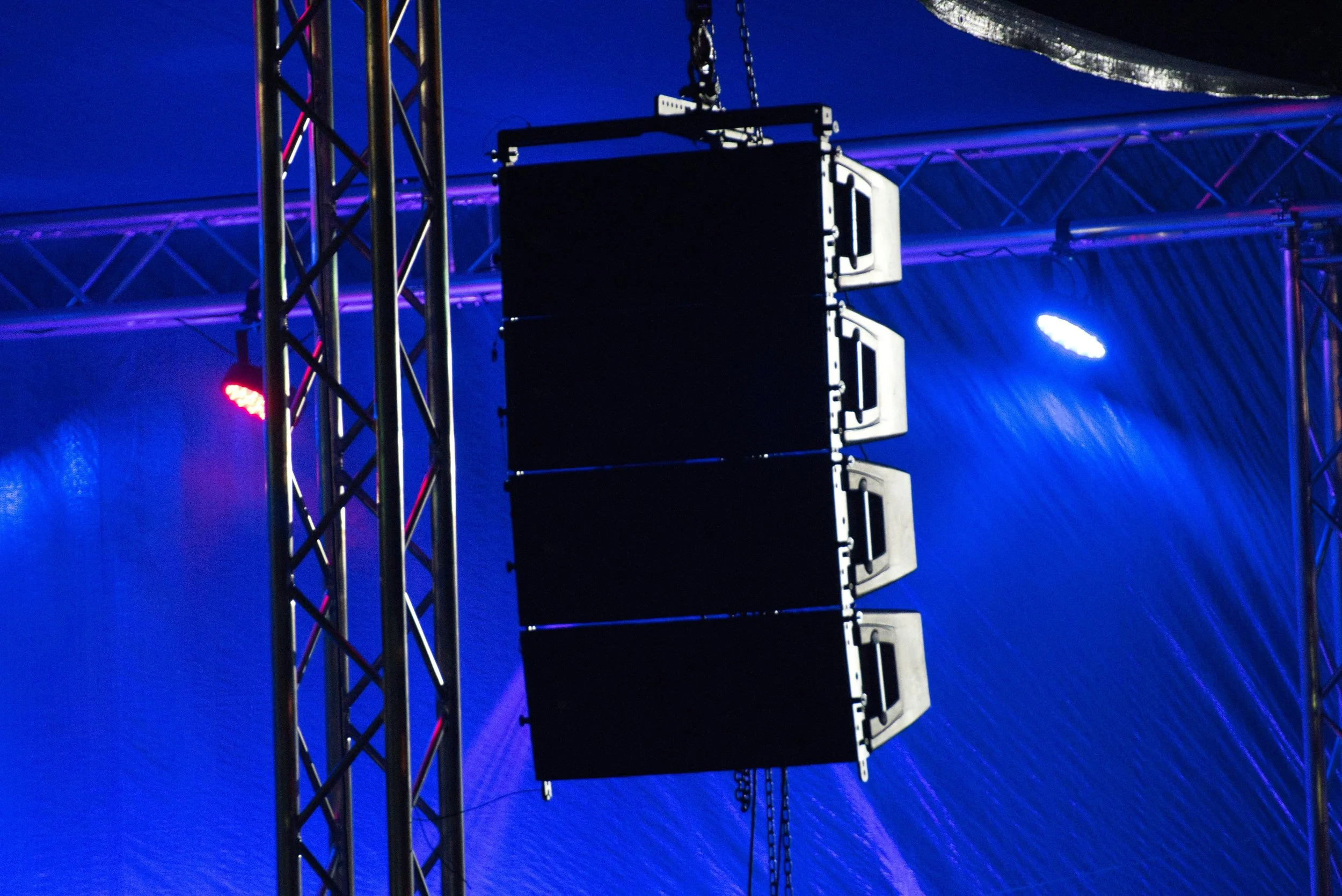 Stage lighting setup with vertical truss structures, colorful lights, and stacked speaker arrays against a dark background.