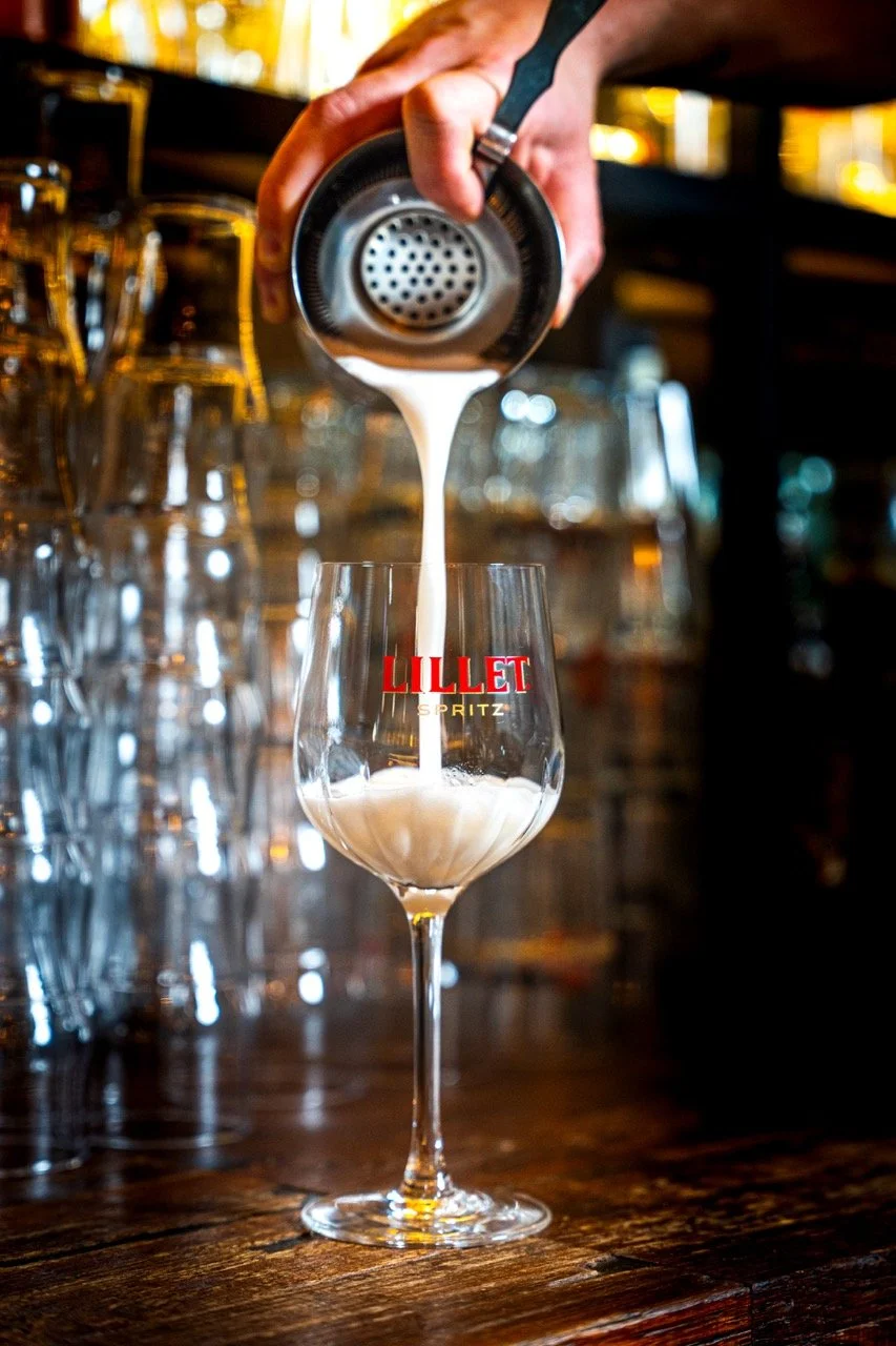 Une main verse une boisson blanche dans un verre à vin gravé avec le logo 'LILLET SPRITZ'.
