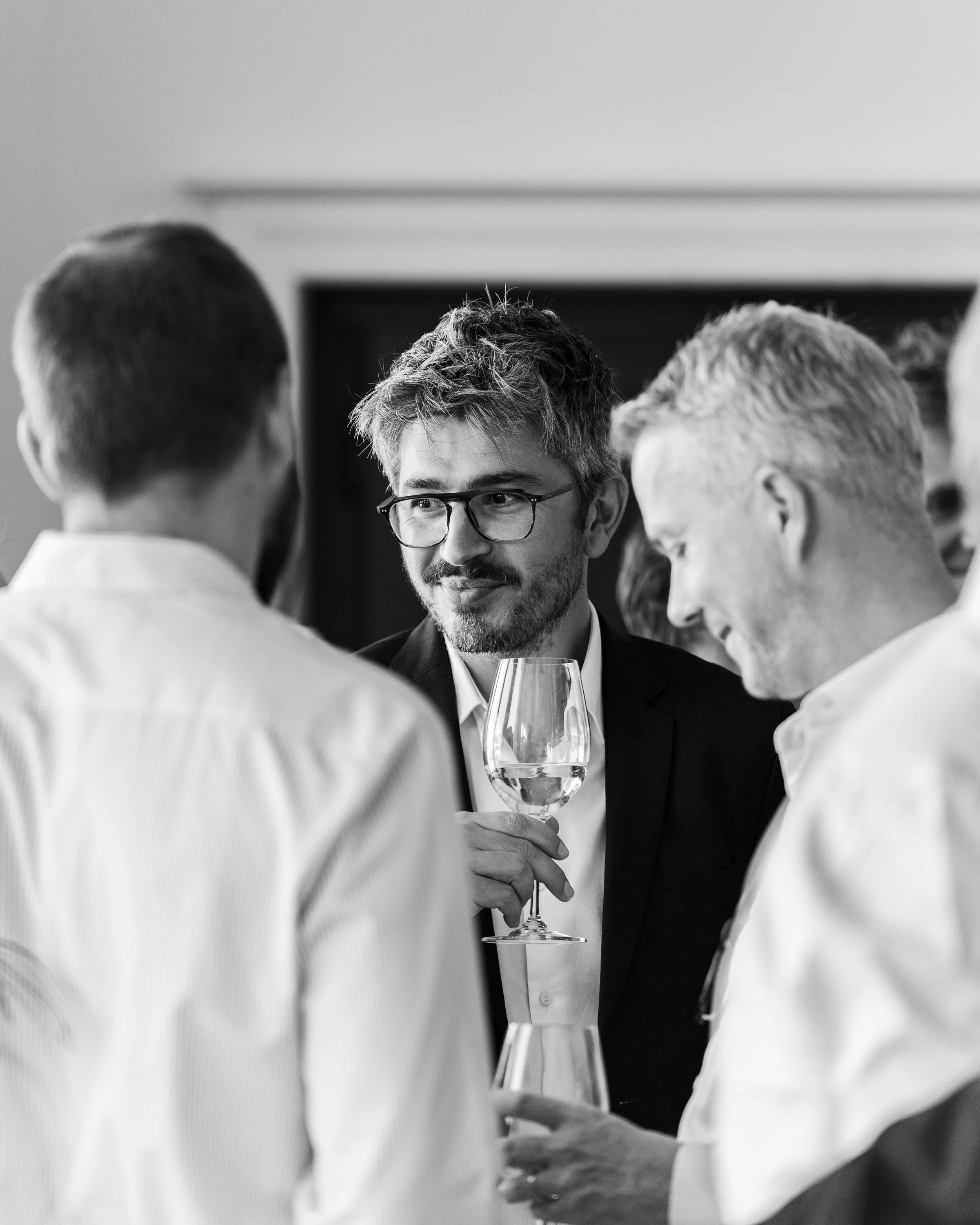 Groupe de personnes en conversation, scène en noir et blanc, un homme avec lunettes tient un verre de vin, entouré d'autres hommes dont certains ont des cheveux gris, dans un cadre intérieur.