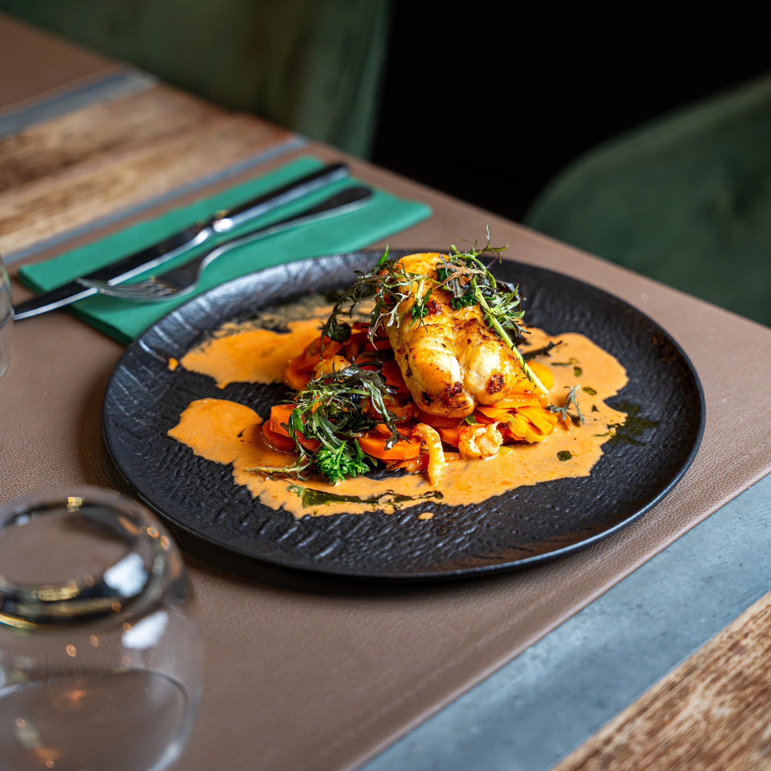 Une assiette noire contenant une viande, des légumes et une sauce, placée sur une table en bois avec des couverts à gauche, dans un environnement intérieur.
