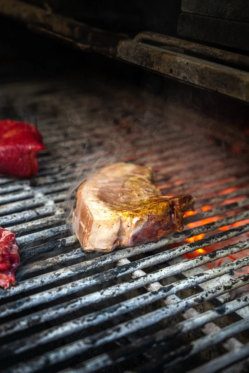Une côte de bœuf en train de cuire sur un barbecue à charbon, avec de la fumée, et d'autres morceaux de viande en arrière-plan.