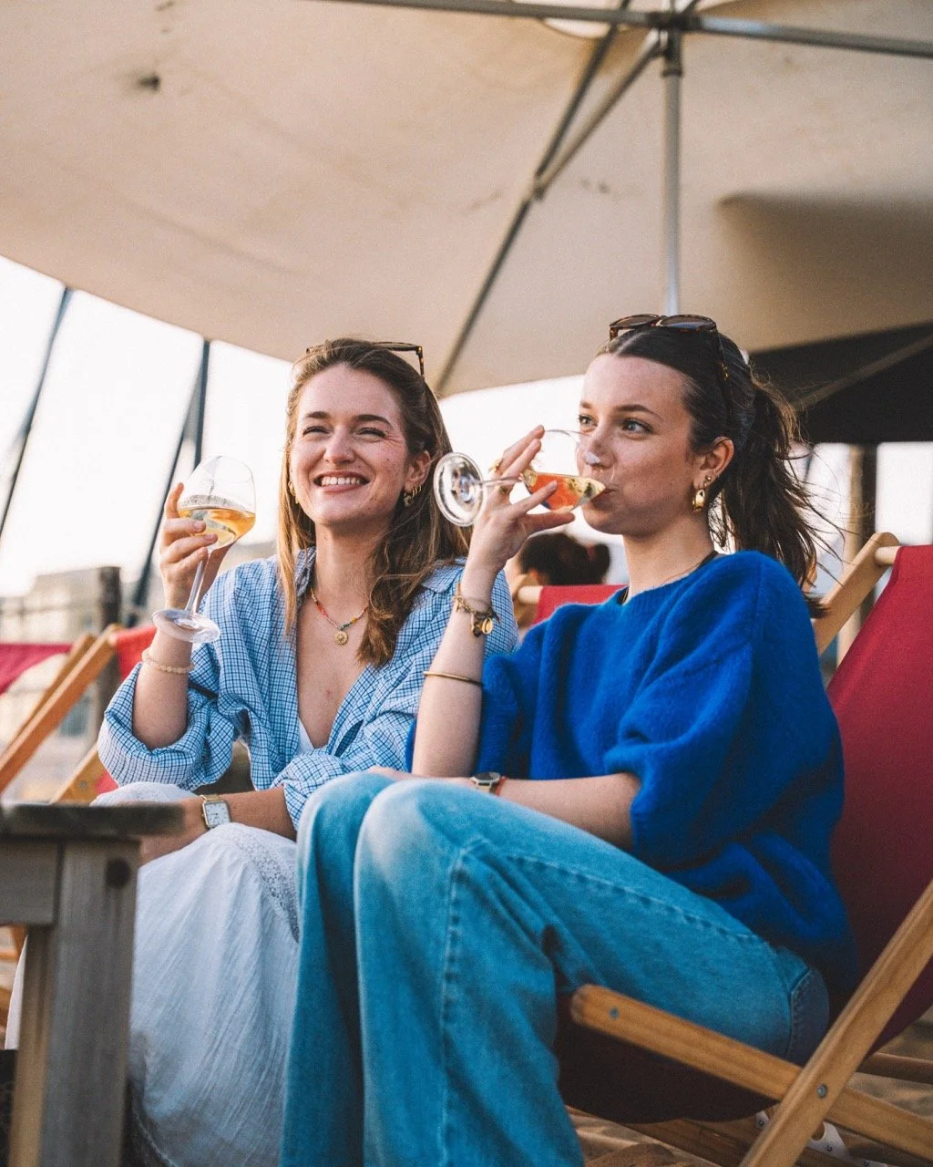 Deux jeunes femmes qui sourient et portent des verres d'alcool sous un parasol, assises sur des chaises longues en plein air.