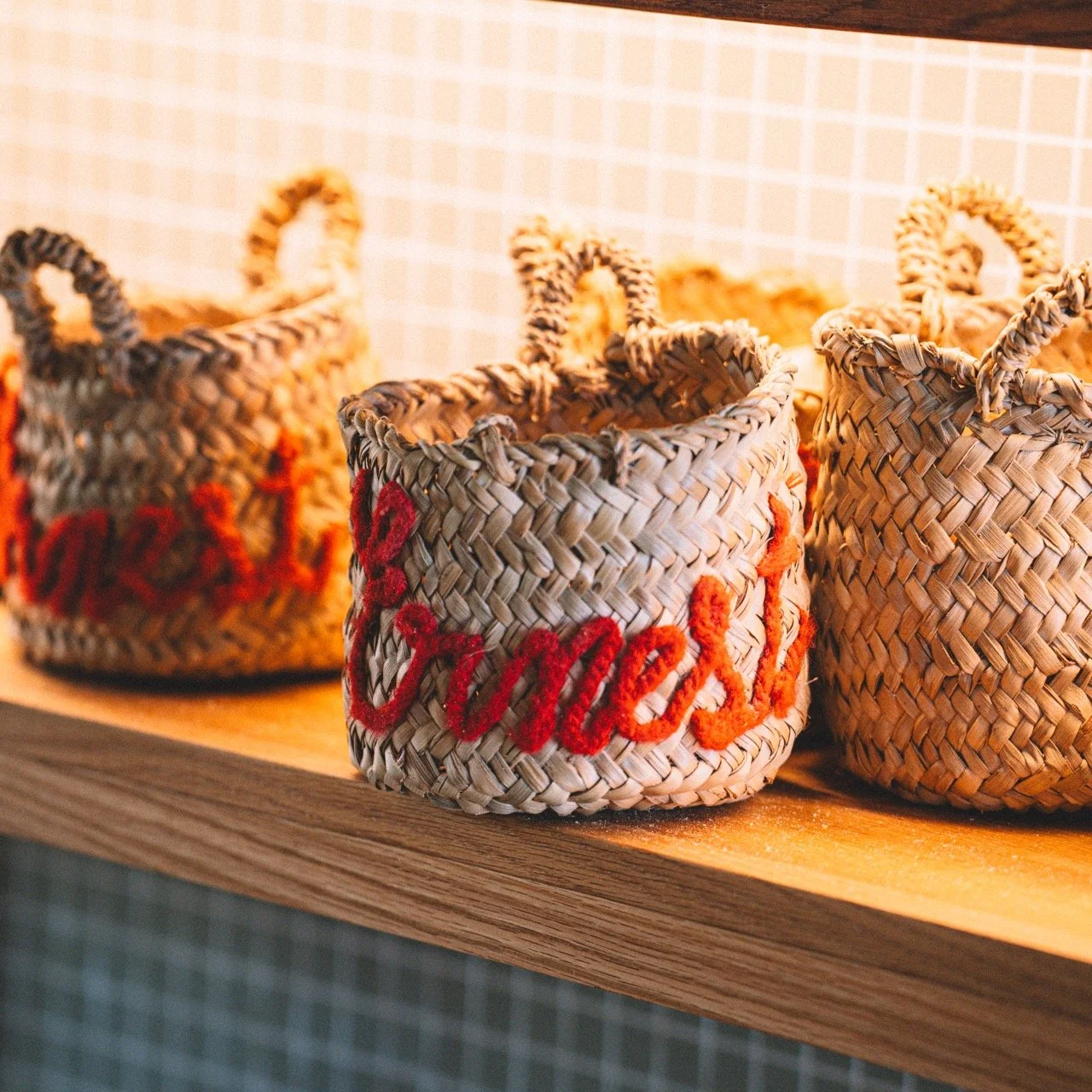 Petites paniers en osier tressé avec poignées, décorés de broderie rouge et orange, posés sur une étagère en bois, fond de mur en carreaux