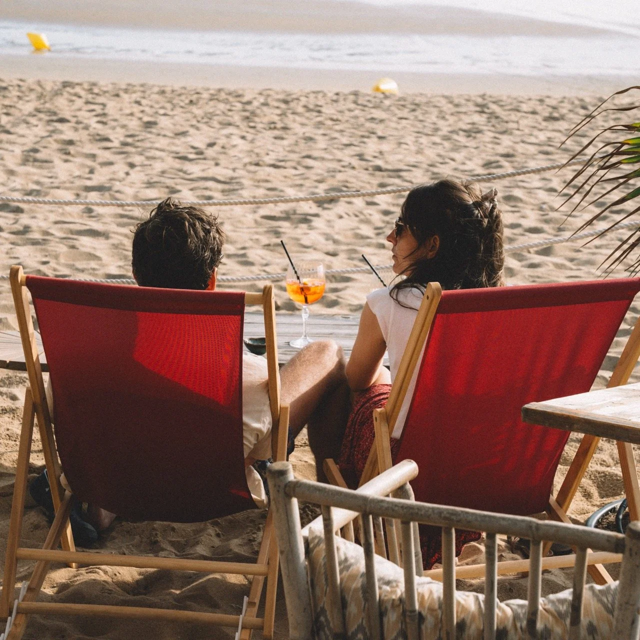Deux personnes assises sur des chaises rouges en terrasse de plage, buvant un cocktail, regardant la mer.