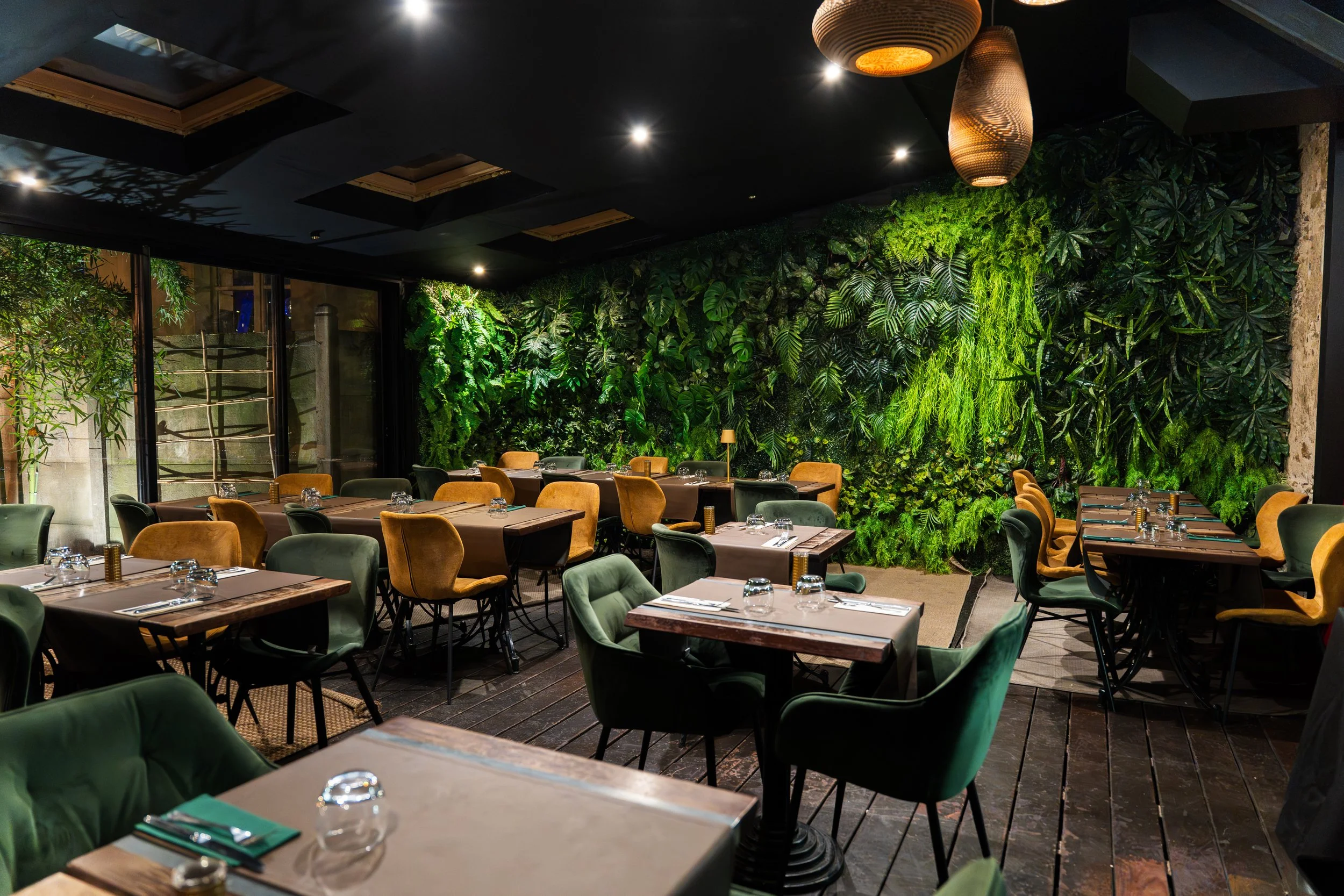 Salle à manger d'un restaurant avec des murs végétaux, des tables en bois, des chaises vertes et jaunes, et un plafond noir orné de suspensions en rotin.