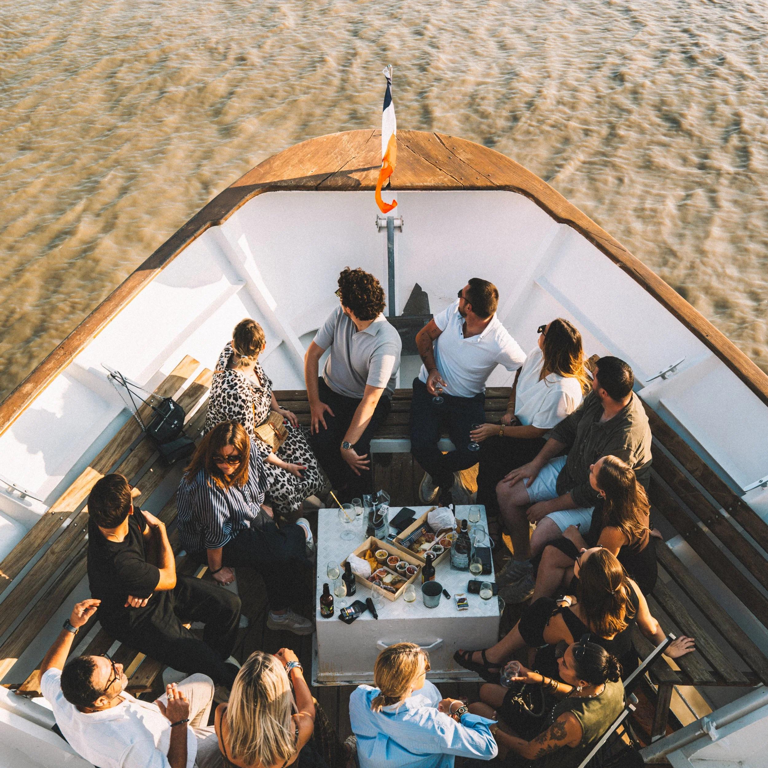 Groupe de personnes assises sur la terrasse d'un bateau profitant d'un repas ou d'une conversation, avec la mer en arrière-plan.