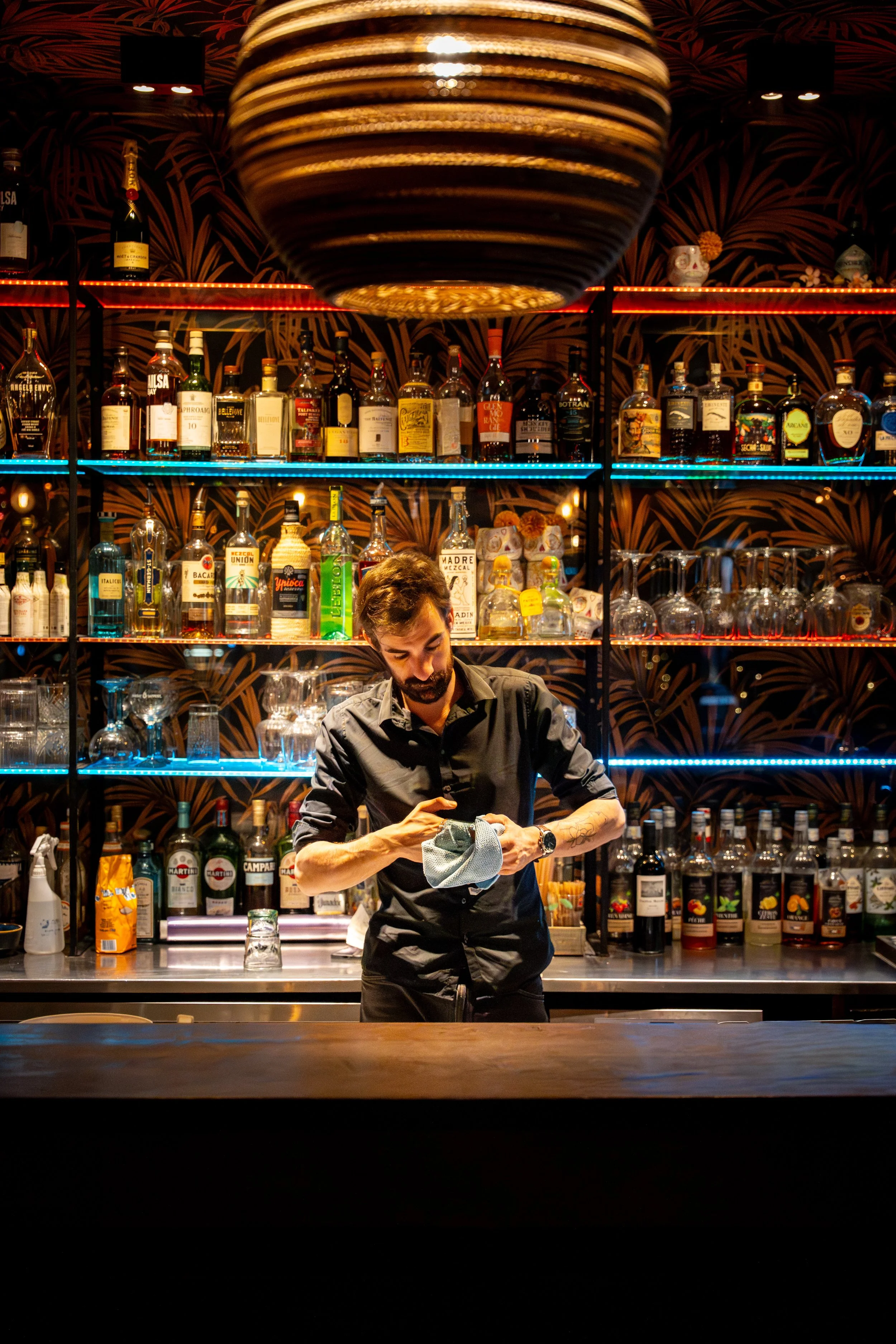Un barman prépare un cocktail derrière un bar avec des étagères remplies de bouteilles d'alcool, sous un éclairage coloré rouge et bleu.