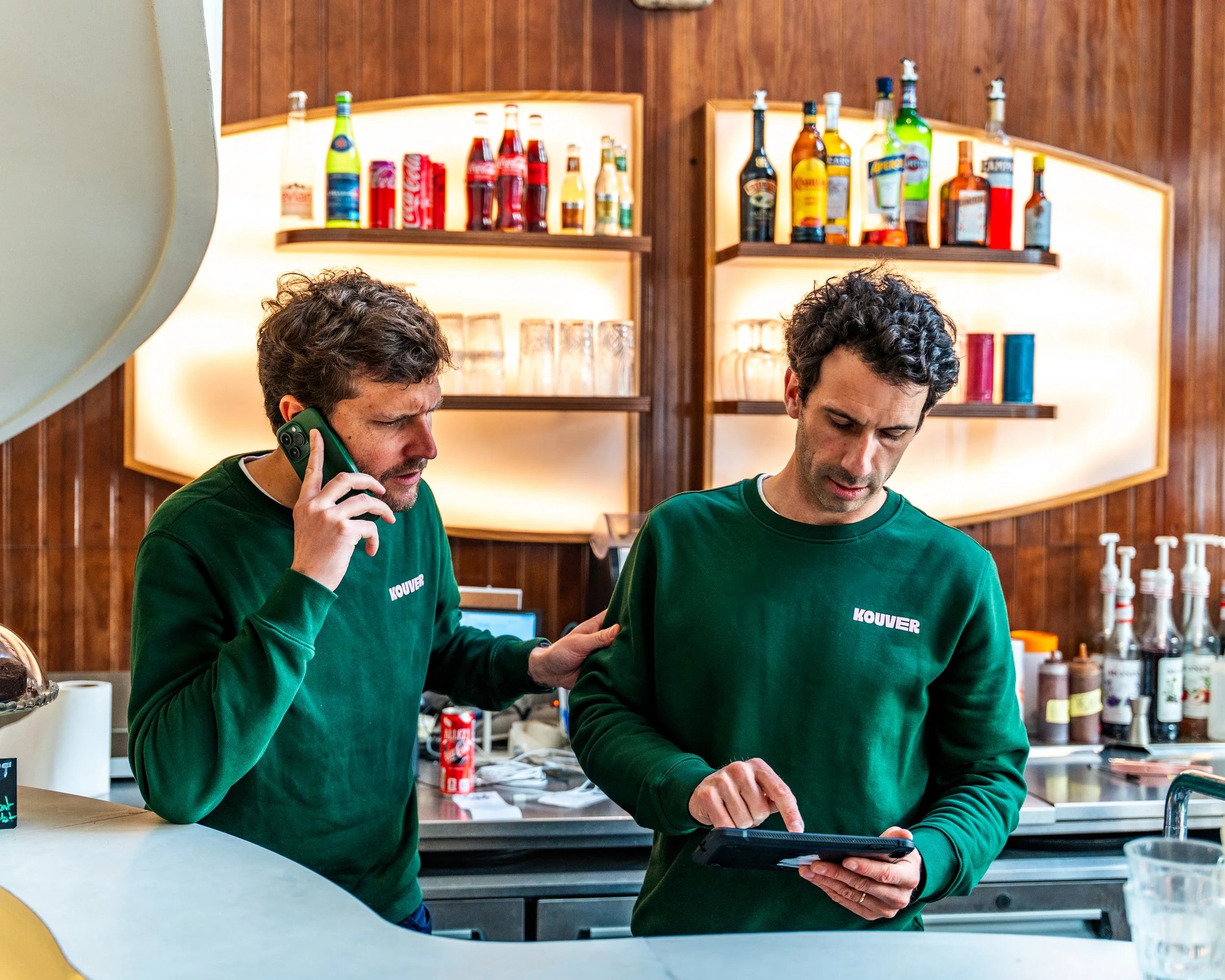 Deux hommes portant des sweat-shirts verts avec le mot 'KOUVER' imprimé, occupés à manipuler un appareil dans une cuisine ou un comptoir de bar avec des étagères en bois derrière eux, contenant diverses bouteilles de boissons alcoolisées et sirops.