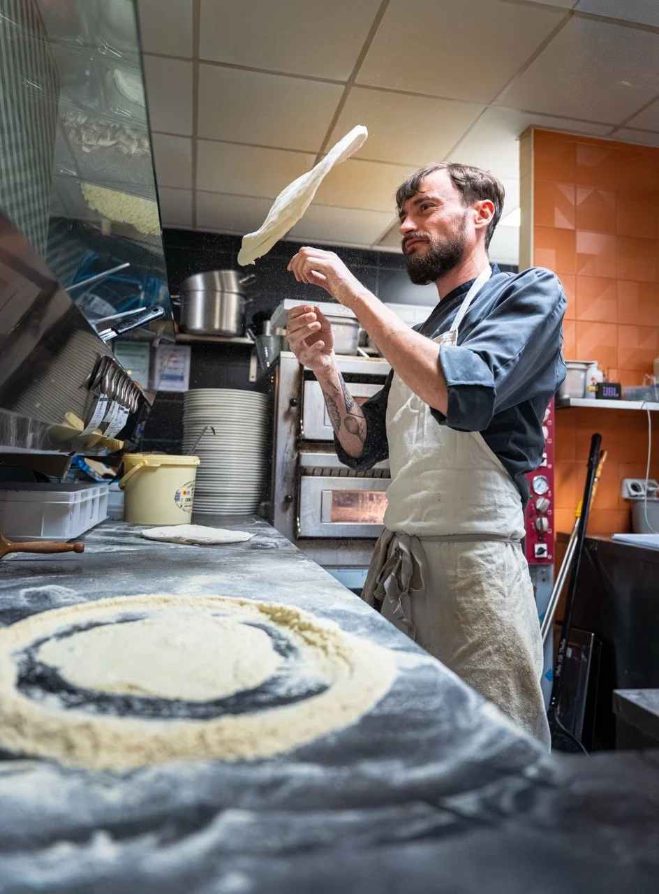 Un chef en cuisine lançant de la pâte à pizza dans l'air, dans un restaurant. La table est recouverte de farine et de pâte, avec divers ustensiles de cuisine en arrière-plan.