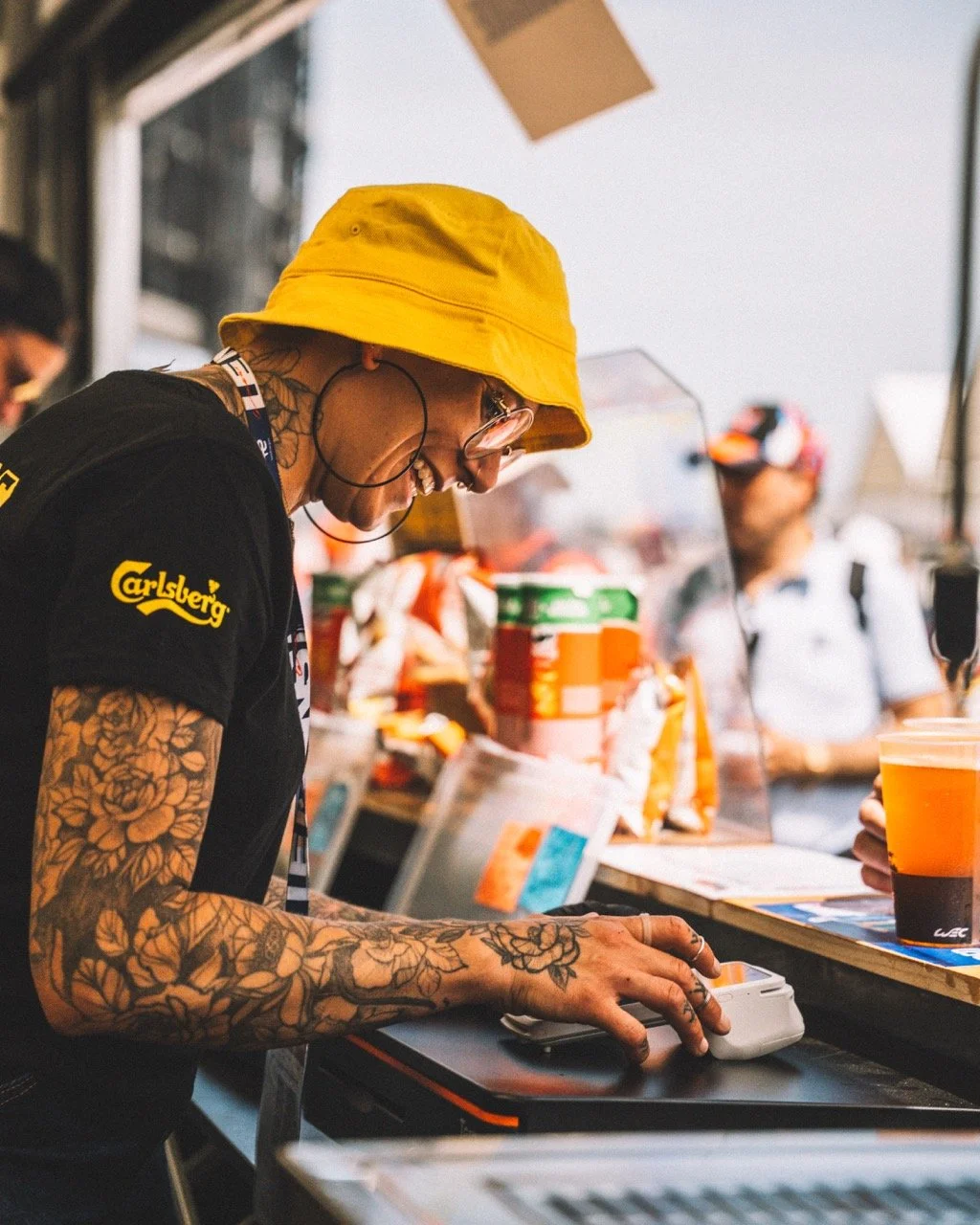 Une personne avec des tatouages, portant un chapeau jaune, des lunettes et un T-shirt noir avec le logo Carlsberg, qui utilise une balance électrique sur un comptoir à une fête ou festival.