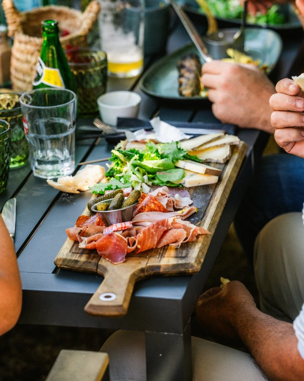 Planche de charcuterie avec jambon, cornichons, fromage, salade, et autres petits plats. Boissons, verres d'eau et de bière sur la table, mains en train de manger.