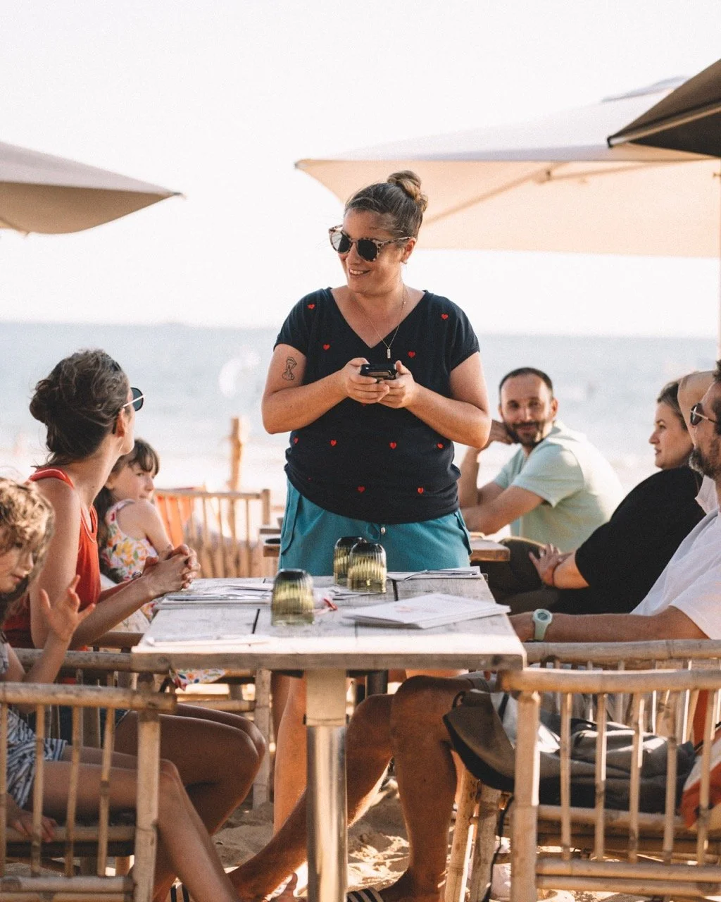 Un groupe de personnes assises autour d'une table de plage, avec une femme en debout tenant un téléphone, sous parasols, à la plage.