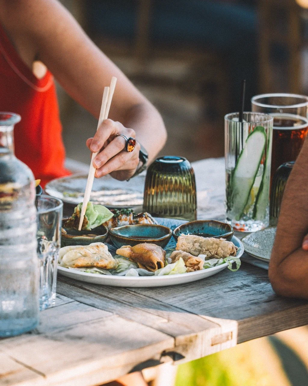 Person utilisant des baguettes pour manger dans un restaurant asiatique, avec un plat de sushi, des boissons comme de l'eau avec du concombre et une boisson sombre, sur une table en bois rustique.