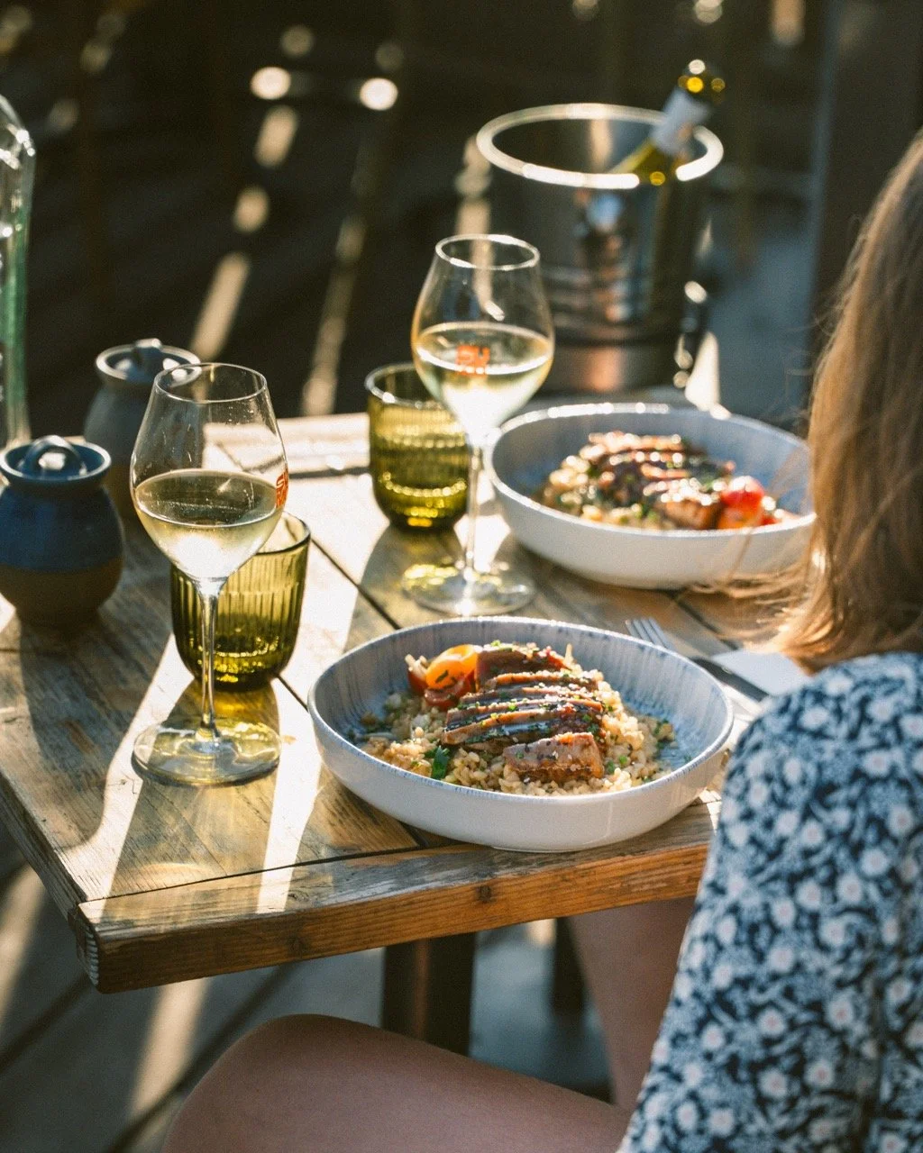 Personne assise à une table en bois avec des plats de riz, légumes et viande, accompagnée de verres de vin blanc, sur fond extérieur en soirée