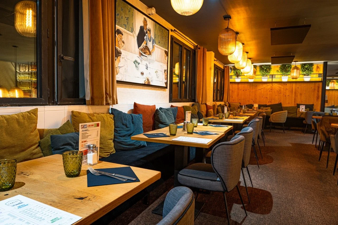 Vue intérieure d'un restaurant chaleureusement décoré avec des tables en bois, des chaises et des coussins colorés, éclairé par des lampes suspendues et décorations végétales.