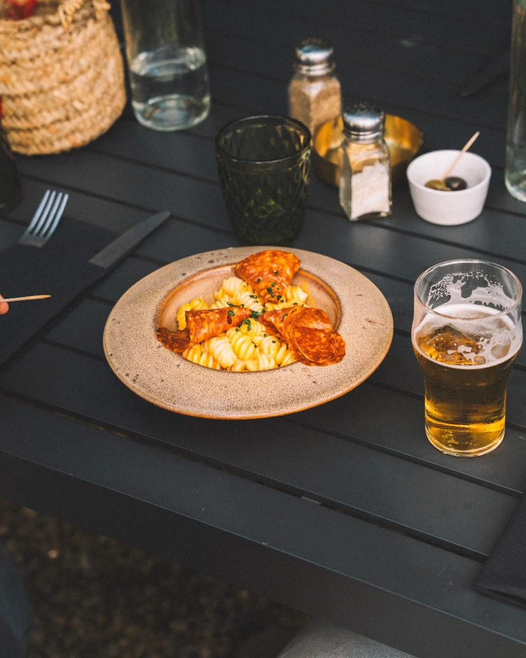 Assiette de pâtes avec de la pizza garnie de pepperoni, servie avec un verre de bière, sur une table en bois noir, entourée de condiments, d'une bouteille d'eau, et d'un petit bol d'olives