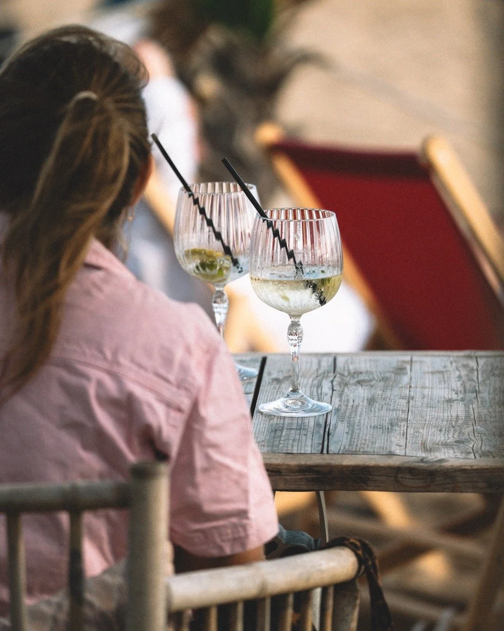 Une personne vue de dos avec des cheveux bruns portant une chemise rose clair, assise à une table en bois avec deux grands verres de vin blanc à moitié pleins, garnis de pailles noires, dans un environnement probablement d'un café ou restaurant.