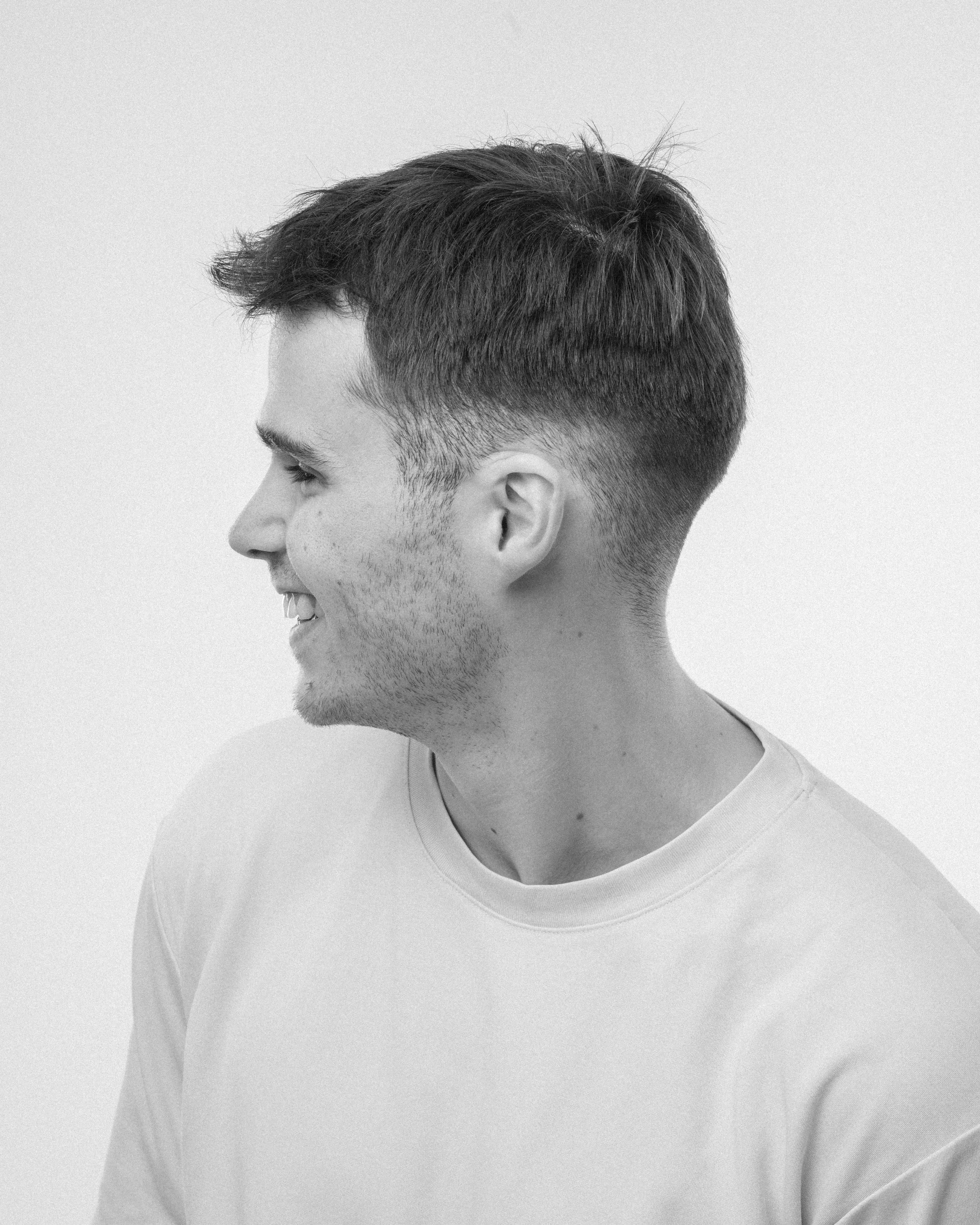 Photo en noir et blanc d'un homme vu en profil, avec une coiffure courte et une barbe légère, portant un t-shirt.