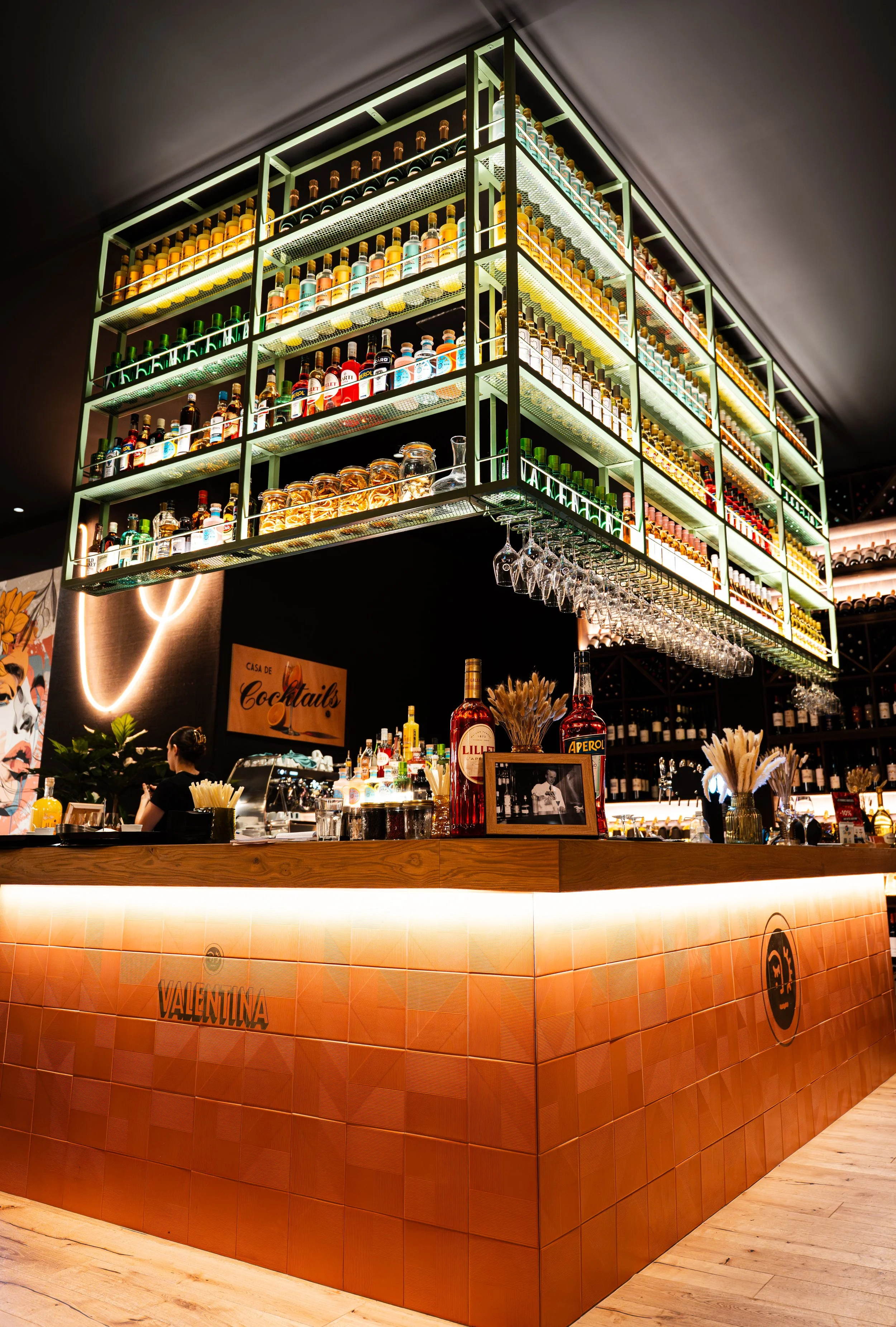 Bar avec étagères lumineuses remplies de bouteilles d'alcool derrière le comptoir en bois. Un panneau indique 'Casa de Cocktails'. Décor moderne avec des fleurs sèches et des verres suspendus.