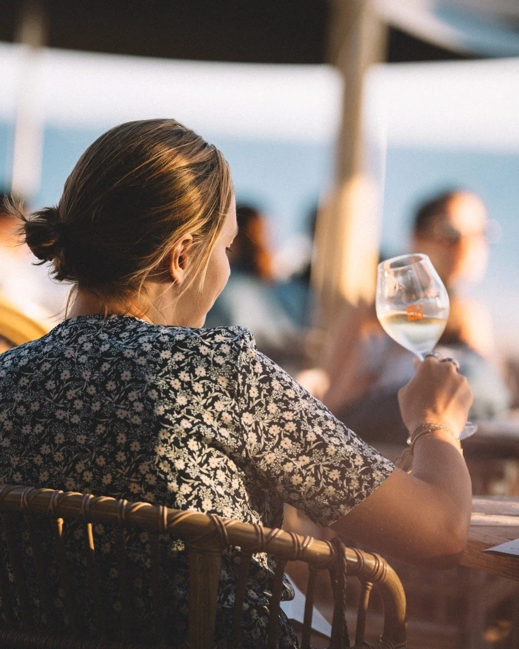 Une femme assise à une terrasse de café ou restaurant, tenant un verre de vin blanc, avec une vue de l'océan au coucher du soleil.