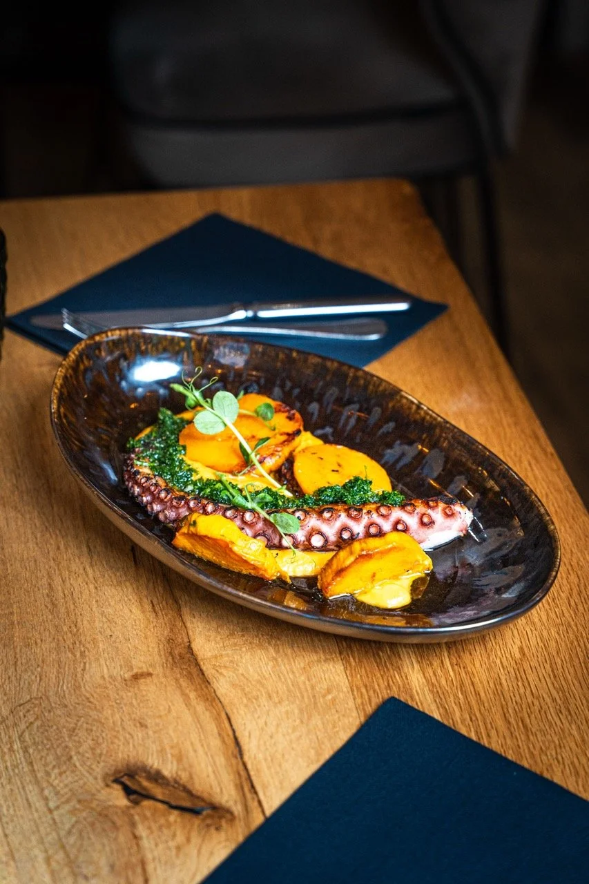 Plat de poulpe avec pommes de terre, épinards et garnitures vertes sur une assiette sombre, posé sur une table en bois.