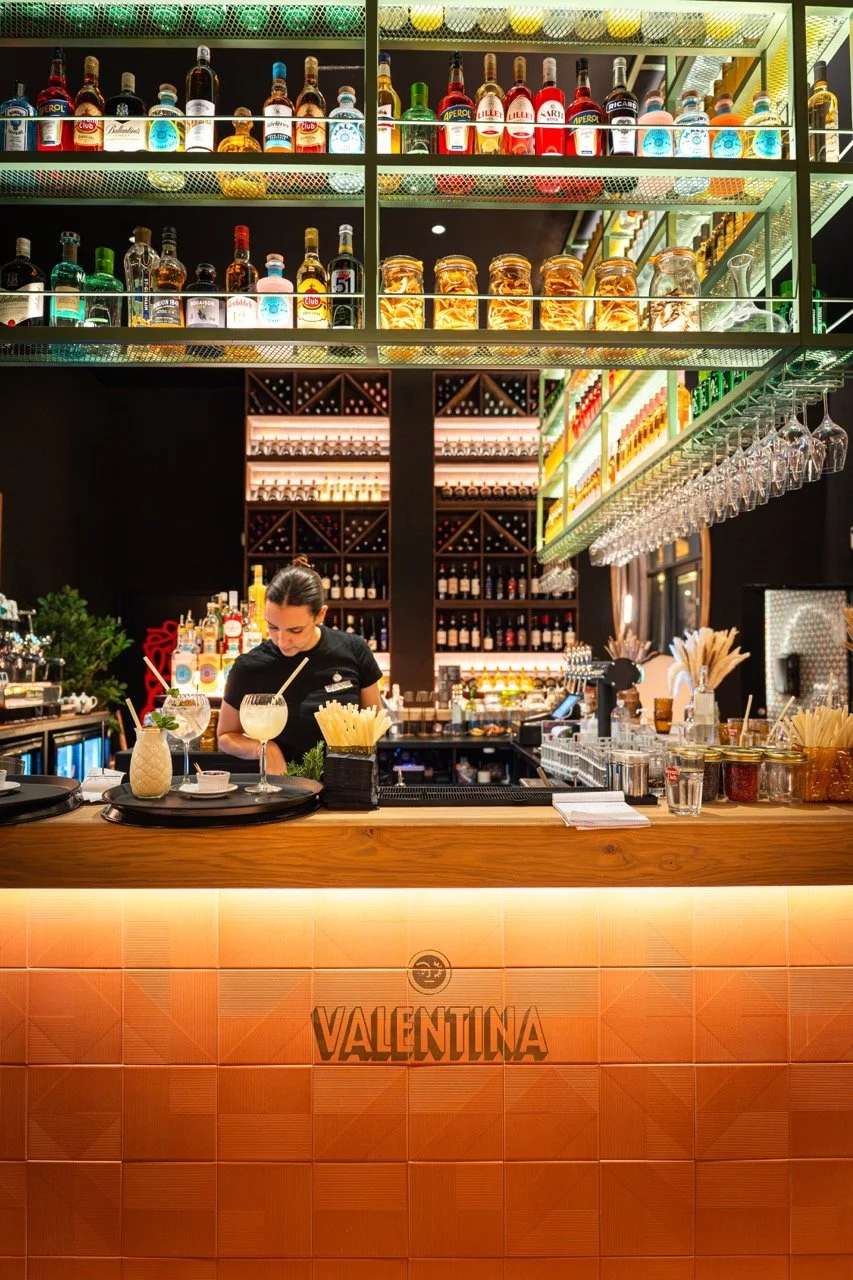 Un bar moderne avec un barman préparant des cocktails, étagères de bouteilles d'alcool, verres à vin suspendus, et un comptoir en bois avec le nom VALENTINA.