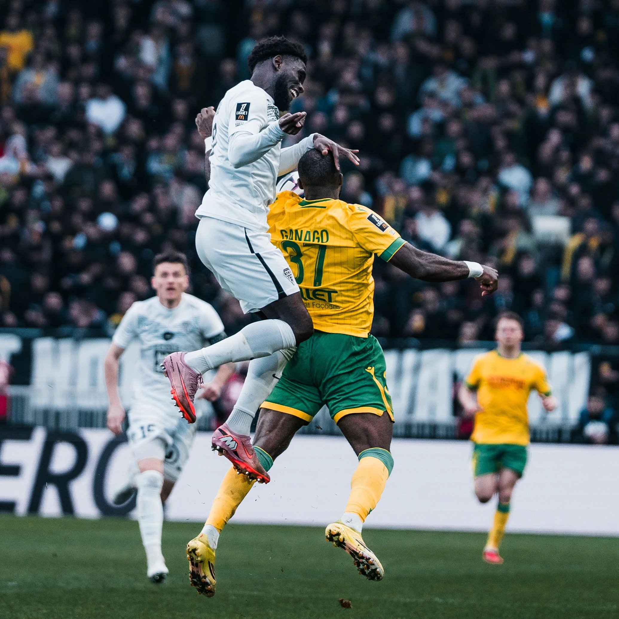 Deux joueurs de football, un en maillot blanc et l'autre en maillot jaune, sautent pour la tête lors d'un match. La foule est visible en arrière-plan.