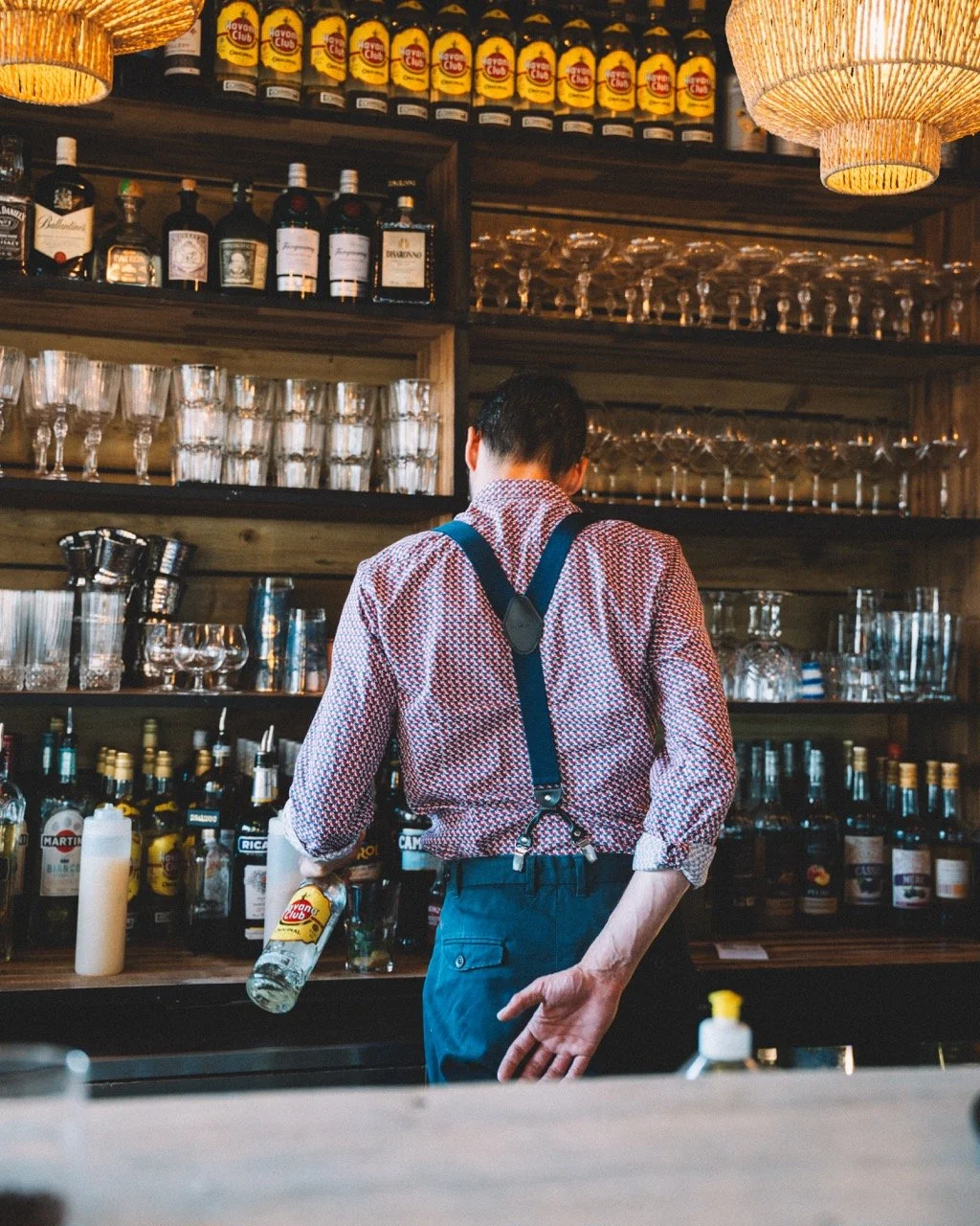 Un homme vu de dos, portant une chemise à motifs rouges et bleus et une salopette, derrière un bar avec diverses bouteilles d'alcool, verres et accessoires de bar, dans un café ou un restaurant.