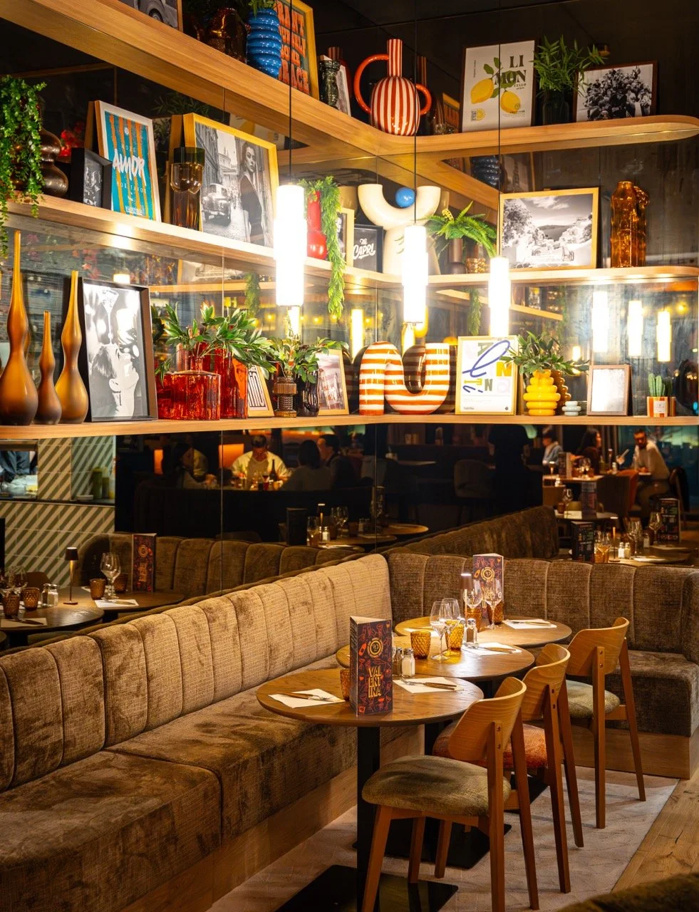 Intérieur d'un restaurant avec des tables en bois, des chaises en bois et une banquette en tissu marron, décorations colorées et étagères avec des vases, des tableaux et des plantes, ambiance chaleureuse.