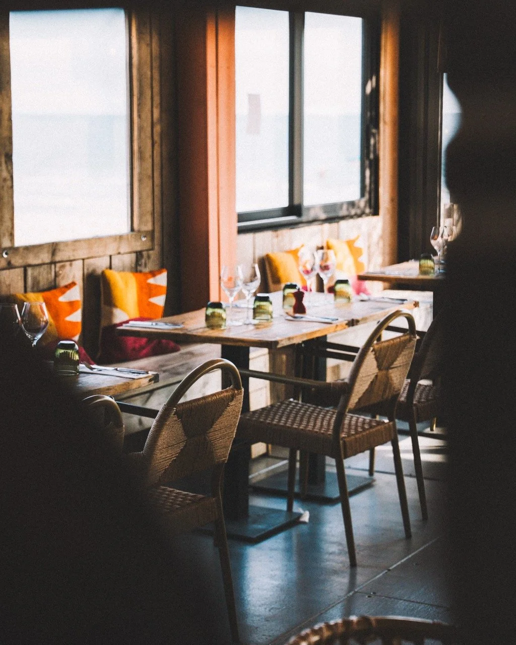 Salle à manger dans un restaurant avec vue sur l'eau, tables avec vaisselle, verres et bougies, décoration en bois, chaises en rotin et coussins colorés