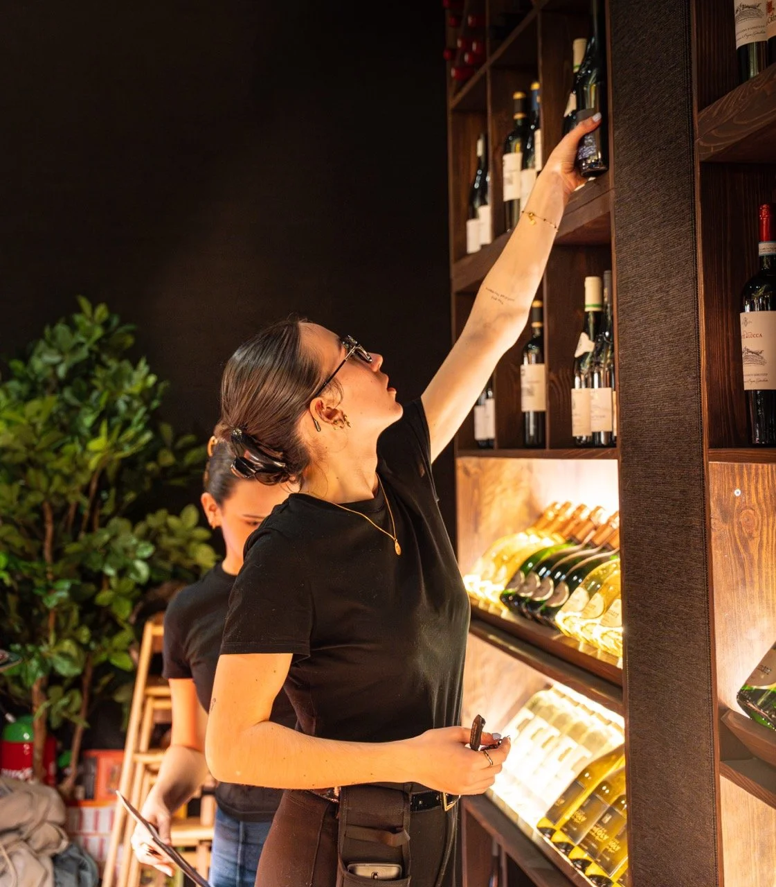 Une femme portant des lunettes cherche une bouteille de vin dans une étagère d'une cave à vin.