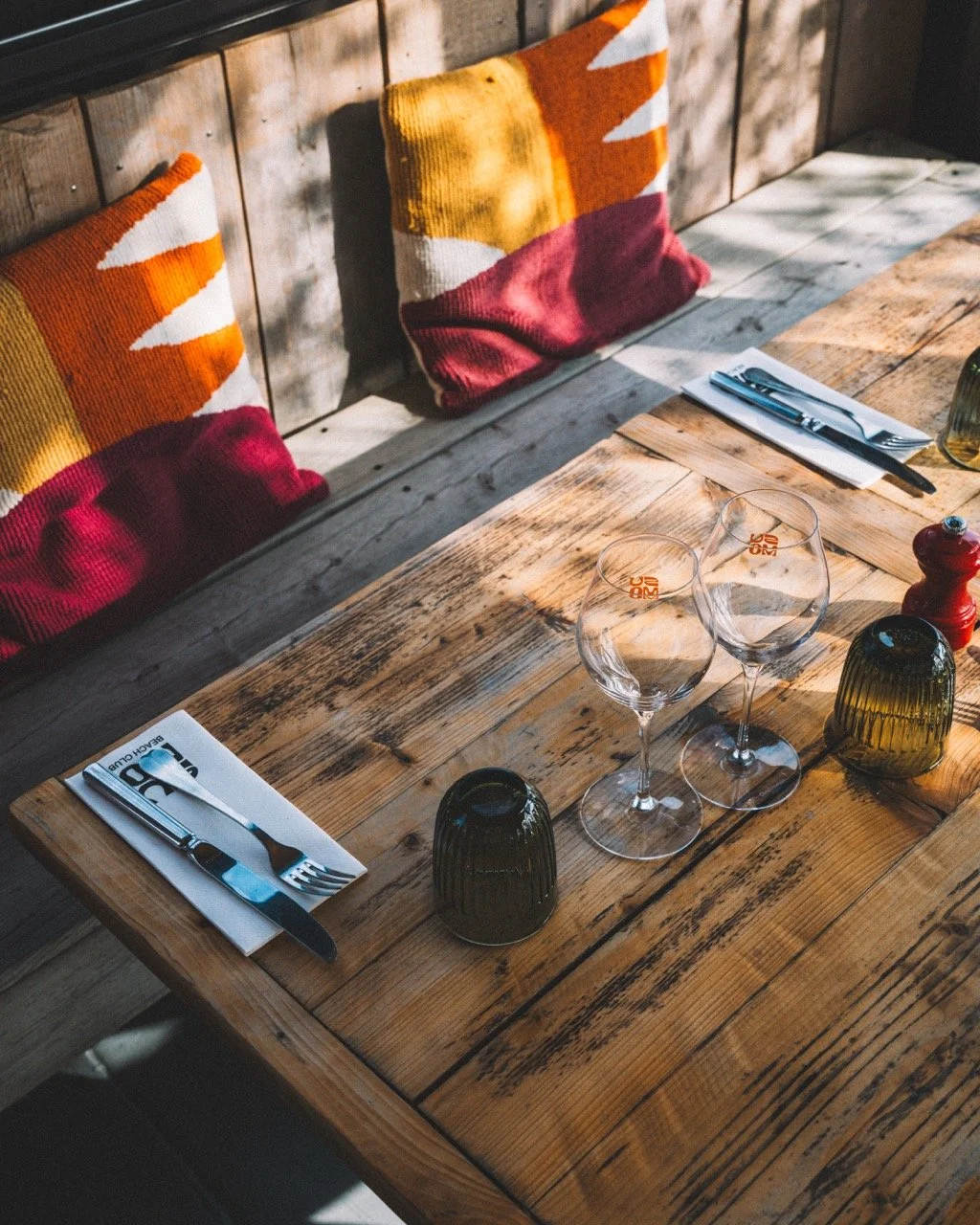 Table en bois dans un restaurant avec deux verres d'eau, deux verres à vin, des fourchettes et couteaux, des napperons avec un logo, deux lampes vertes, un poivre, et des coussins colorés avec des motifs géométriques sur un banc en bois.