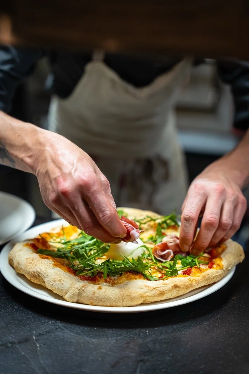 Une personne prépare une pizza avec de la roquette, du jambon, de la mozzarella sur une pâte à pizza.