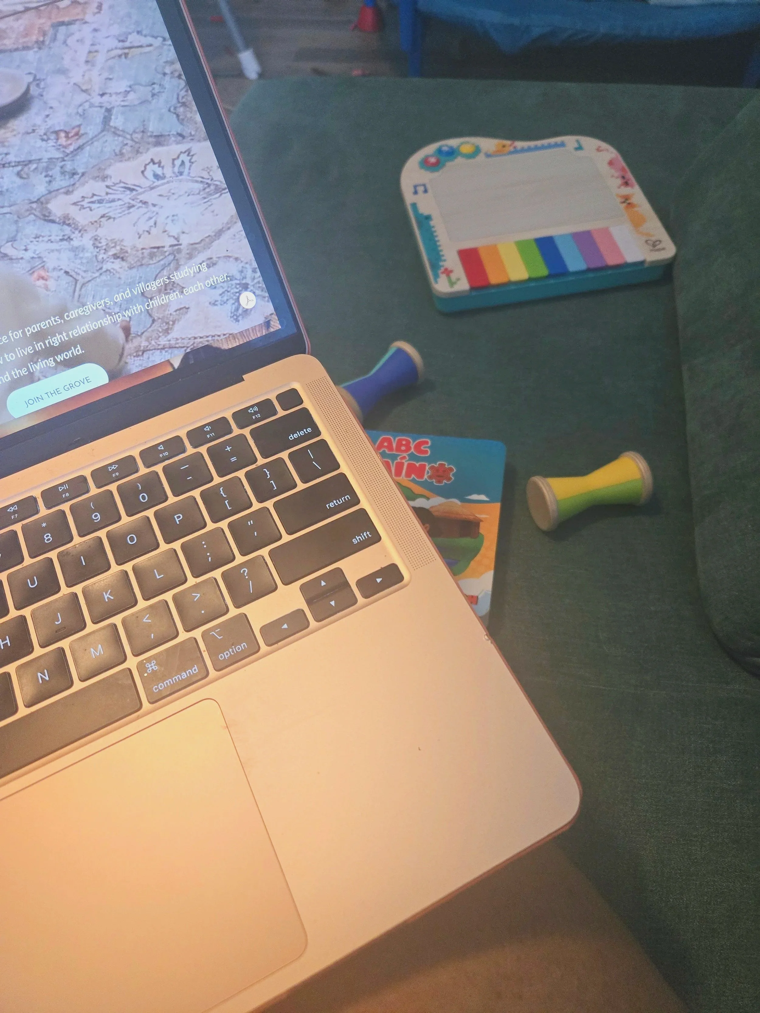 a pink macbook sits on the lap of the person using it who is sitting on a play couch scattered with a toy piano, wooden objects, and a book..