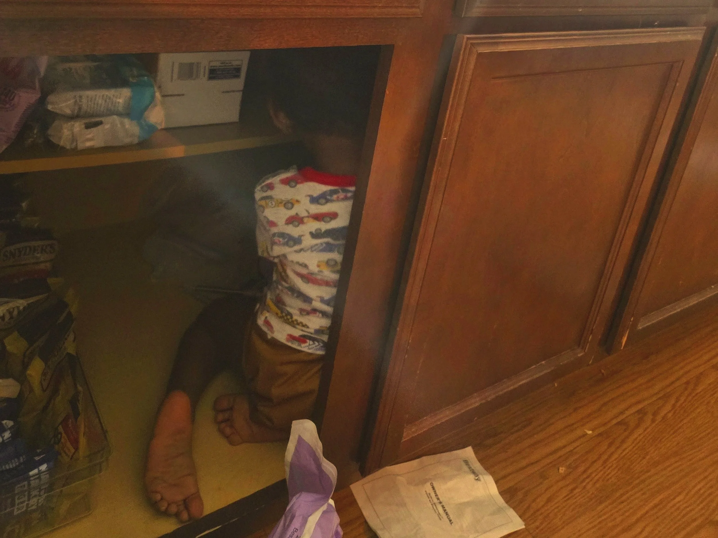 A toddler sitting inside of a lower kitchen cabinet filled with pretzels, seaweed, bars, and popcorn. The child wears a brown cloth diaper and ringer tee with racing cars in primary colors