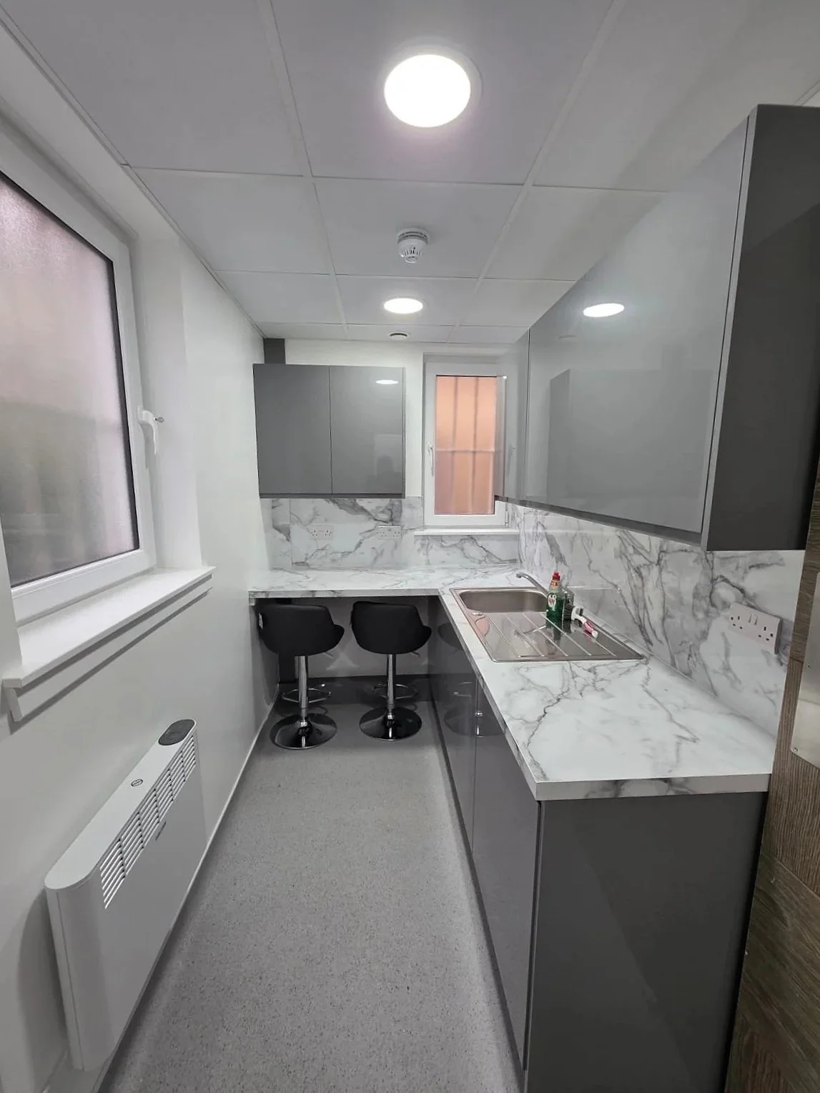 Modern kitchen with gray cabinets, marble countertops, and a marble backsplash, featuring a stainless steel sink with cleaning supplies, two black bar stools at the counter, and a window with frosted glass.
