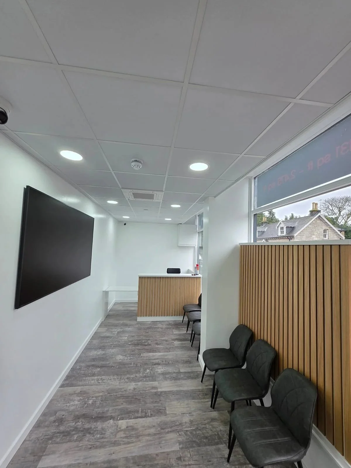 An empty waiting room with black chairs along the wall, a large flat-screen TV, wooden panel accents, and a reception desk near the window.