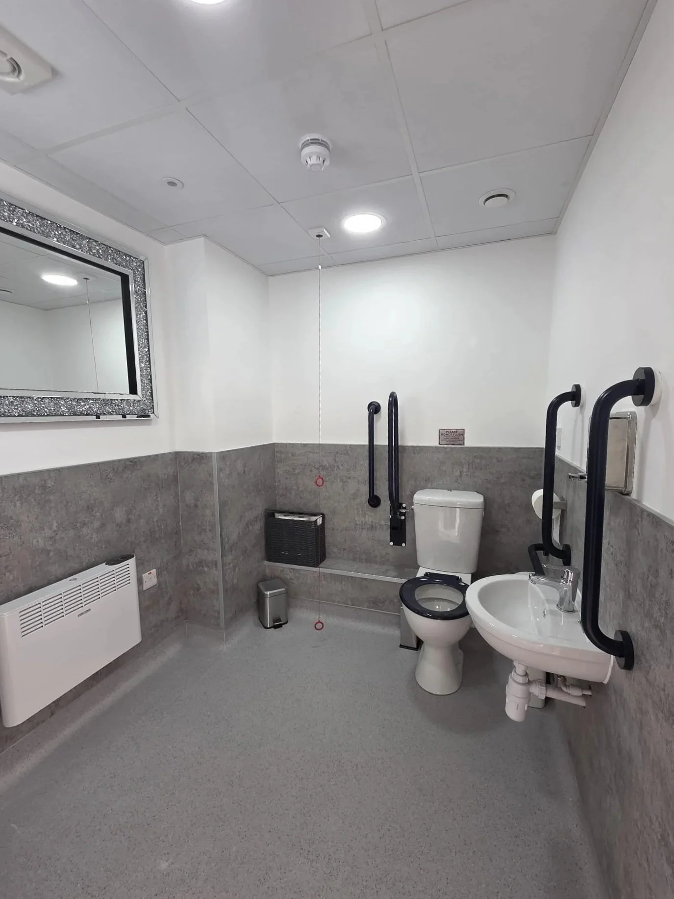 Accessible bathroom with white toilet, white sink, handrails, mirror, heater, and gray flooring.
