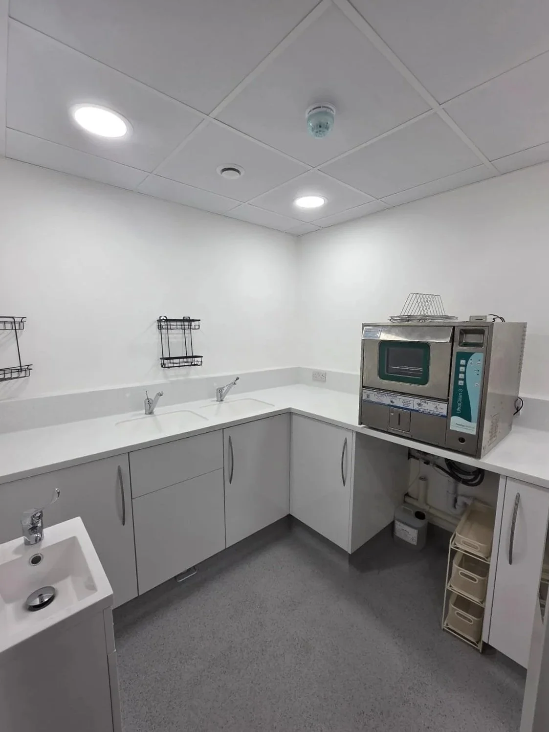Clean medical or laboratory sterilization room with white cabinets, a countertop, a small sink, wire racks on the wall, and an autoclave machine on the countertop.