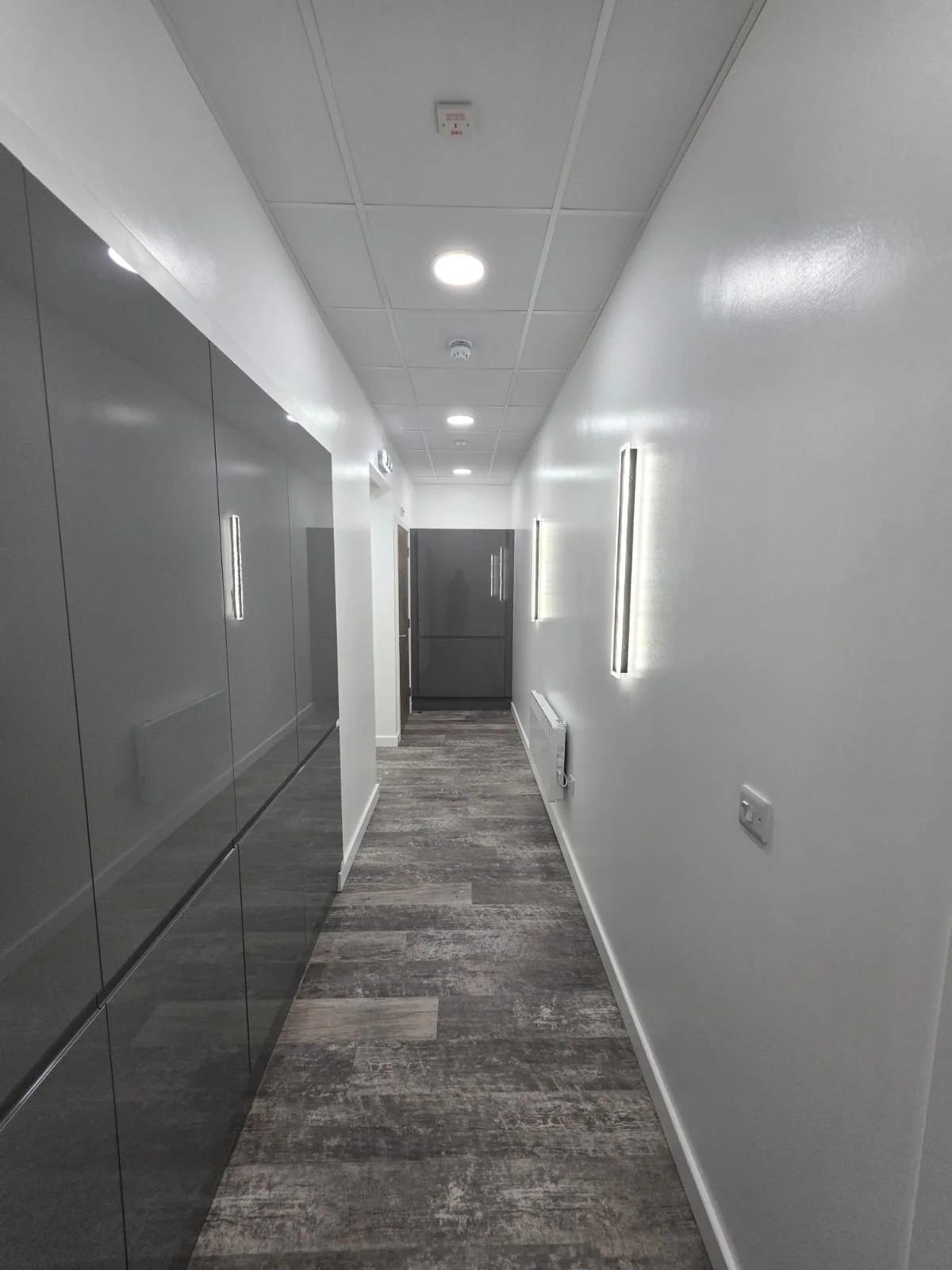 A modern, clean hallway with grey cabinets on the left, white walls, and wood-like flooring. The ceiling has recessed lights and mounted vertical lights along the walls. At the end of the hallway, there is a grey cabinet or wardrobe.