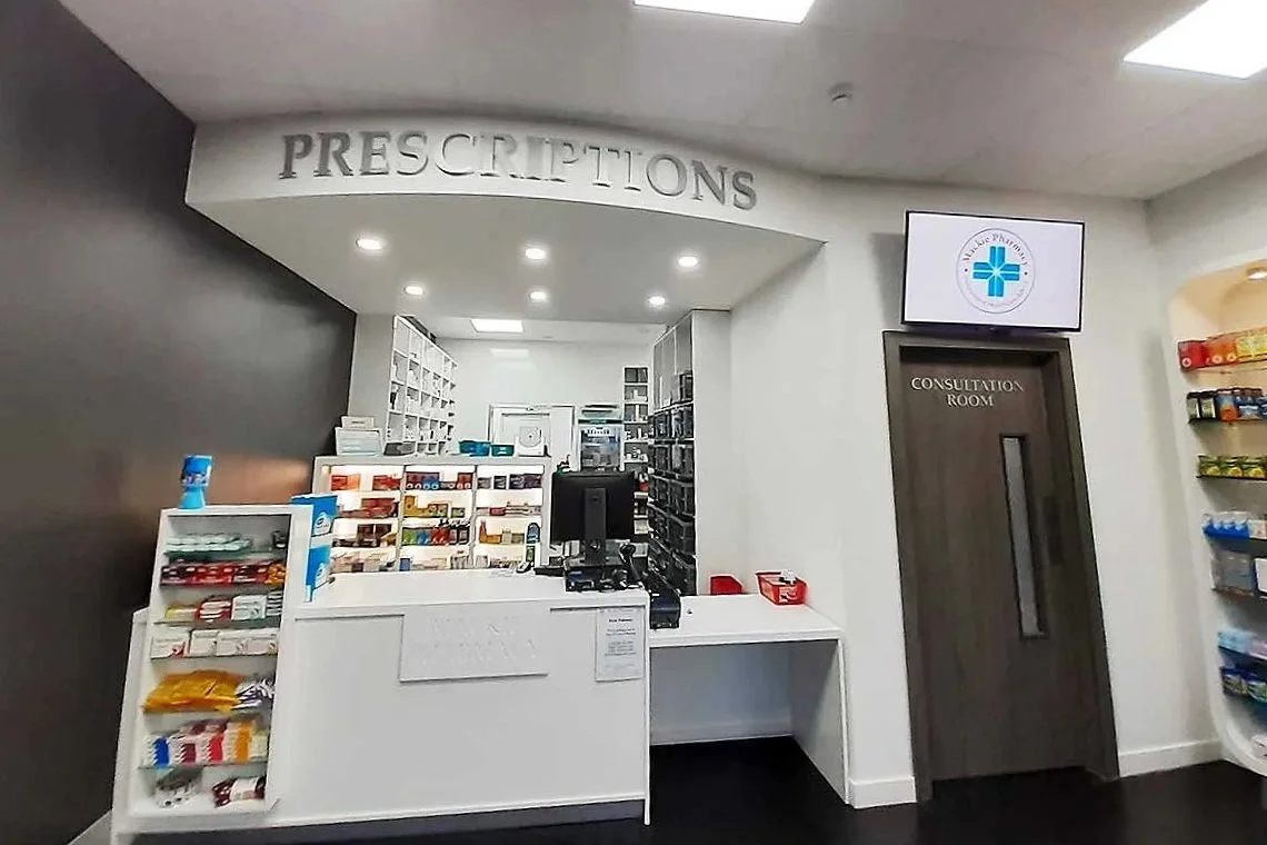 Pharmacy prescription counter with shelves of medications, a computer, and a consultation room door in the background.