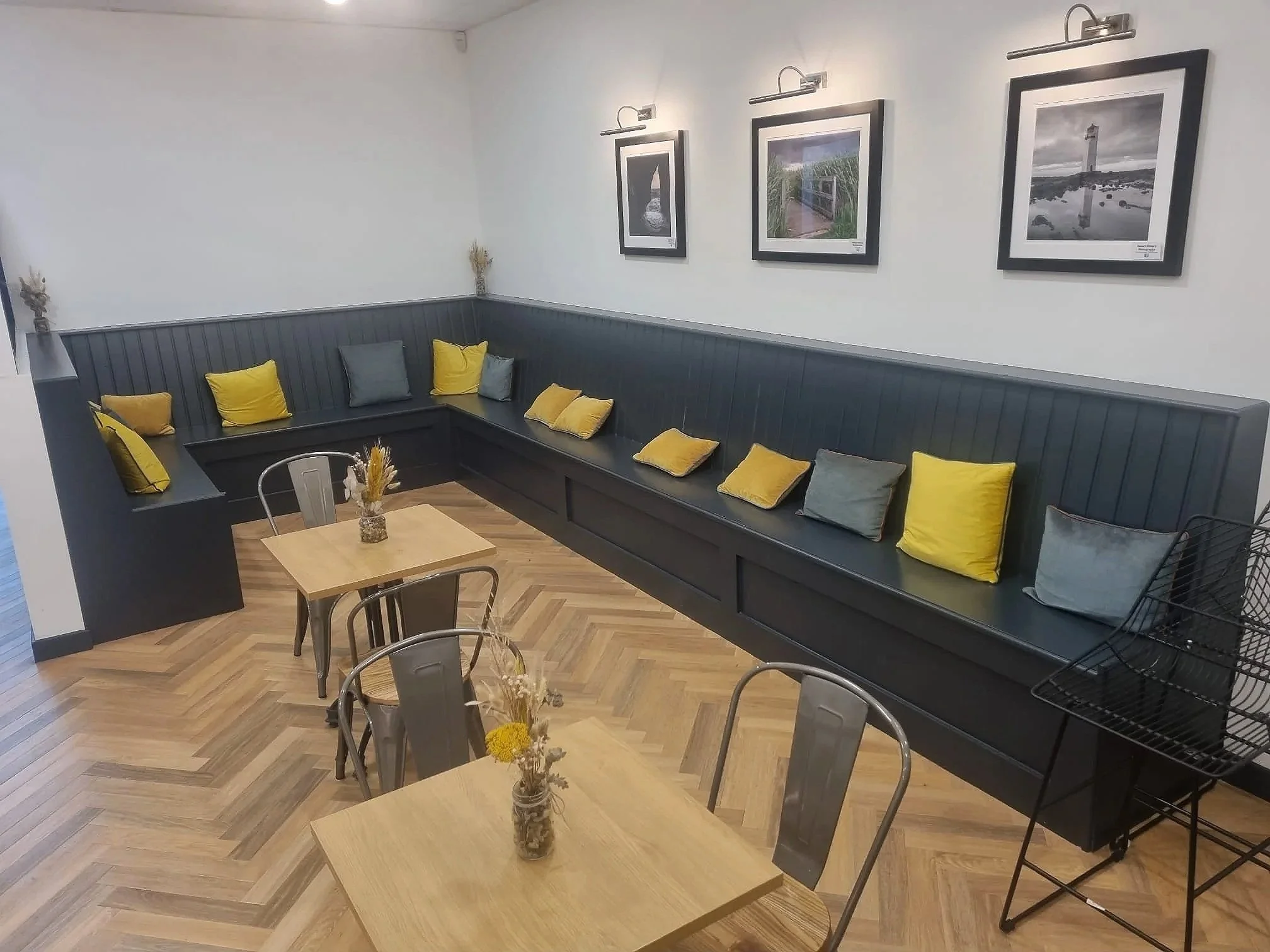 A cozy indoor seating area with a black L-shaped bench accented with yellow and gray pillows. The add-on tables have small vases with dried flowers. The white wall's upper portion is decorated with three framed black-and-white photographs of lighthou