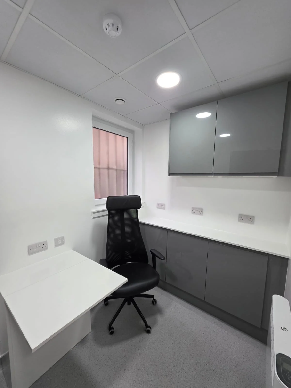 An empty office room with a black swivel chair, white desk, gray cabinets, white walls, a window, and ceiling lights.