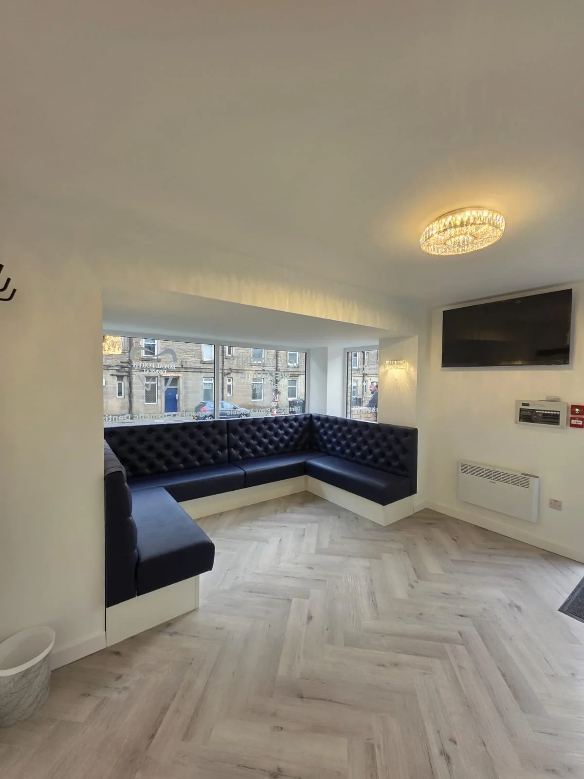 A modern lounge with black tufted leather seating arranged in a corner near large windows, light wooden herringbone floors, a flat-screen TV on the wall, and a decorative light fixture on the ceiling.